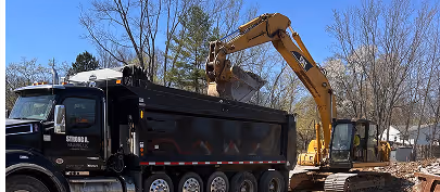 Yellow excavator loading dirt into a black dump truck beside a dirt road with leafless trees in the background.