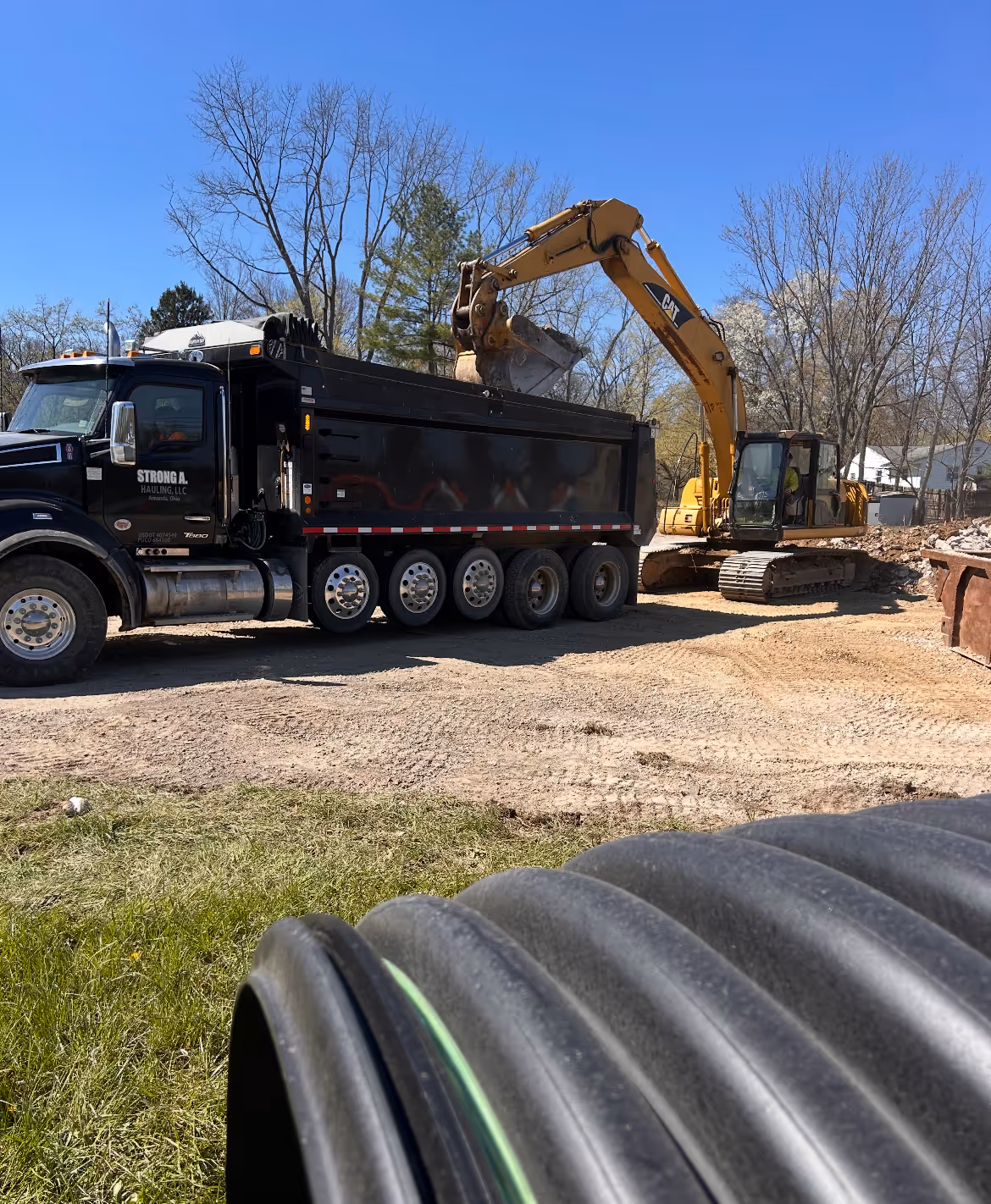 Yellow CAT excavator loading soil into a black Strong A. Hauling dump truck on a dirt construction site with trees in the background.