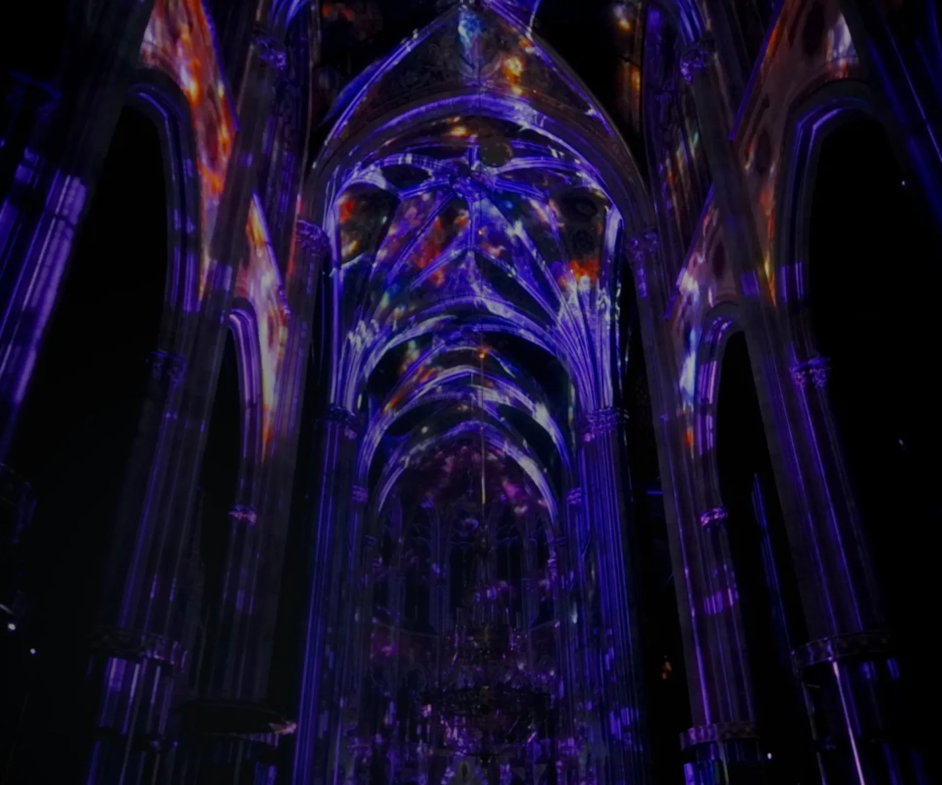 Vivid multicolor light projection illuminating the interior of a Gothic cathedral, with purple, pink, and blue hues highlighting the tall arches, stained glass, and ornate altar.