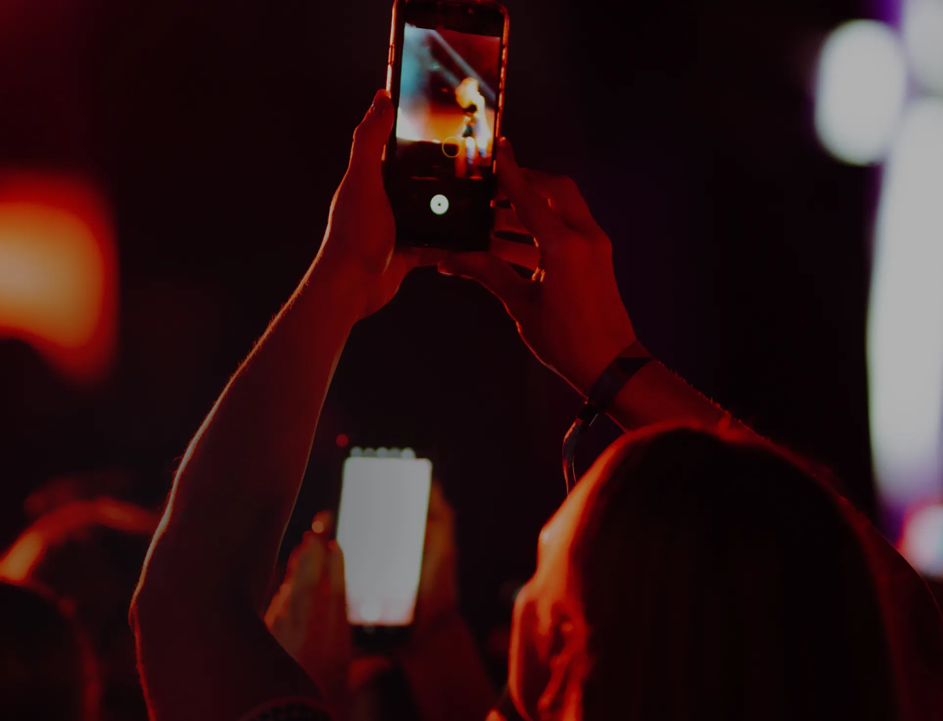 Person holding up a smartphone to record a live performance, surrounded by warm stage lighting and an audience.