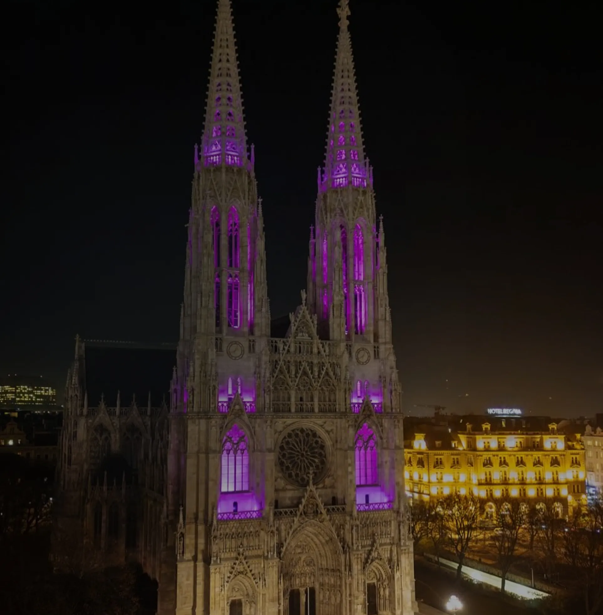 Night view of a Gothic cathedral illuminated with purple lights, with city buildings glowing in the background.”