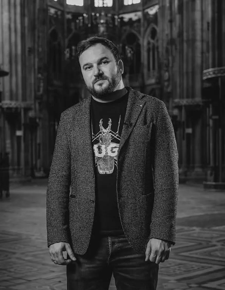 Black and white portrait of a man standing in a grand cathedral interior, wearing a textured blazer and a printed T-shirt.