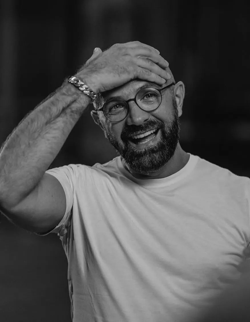 Black and white portrait of a smiling man wearing glasses and a white T-shirt, touching his head with one hand in a candid moment.