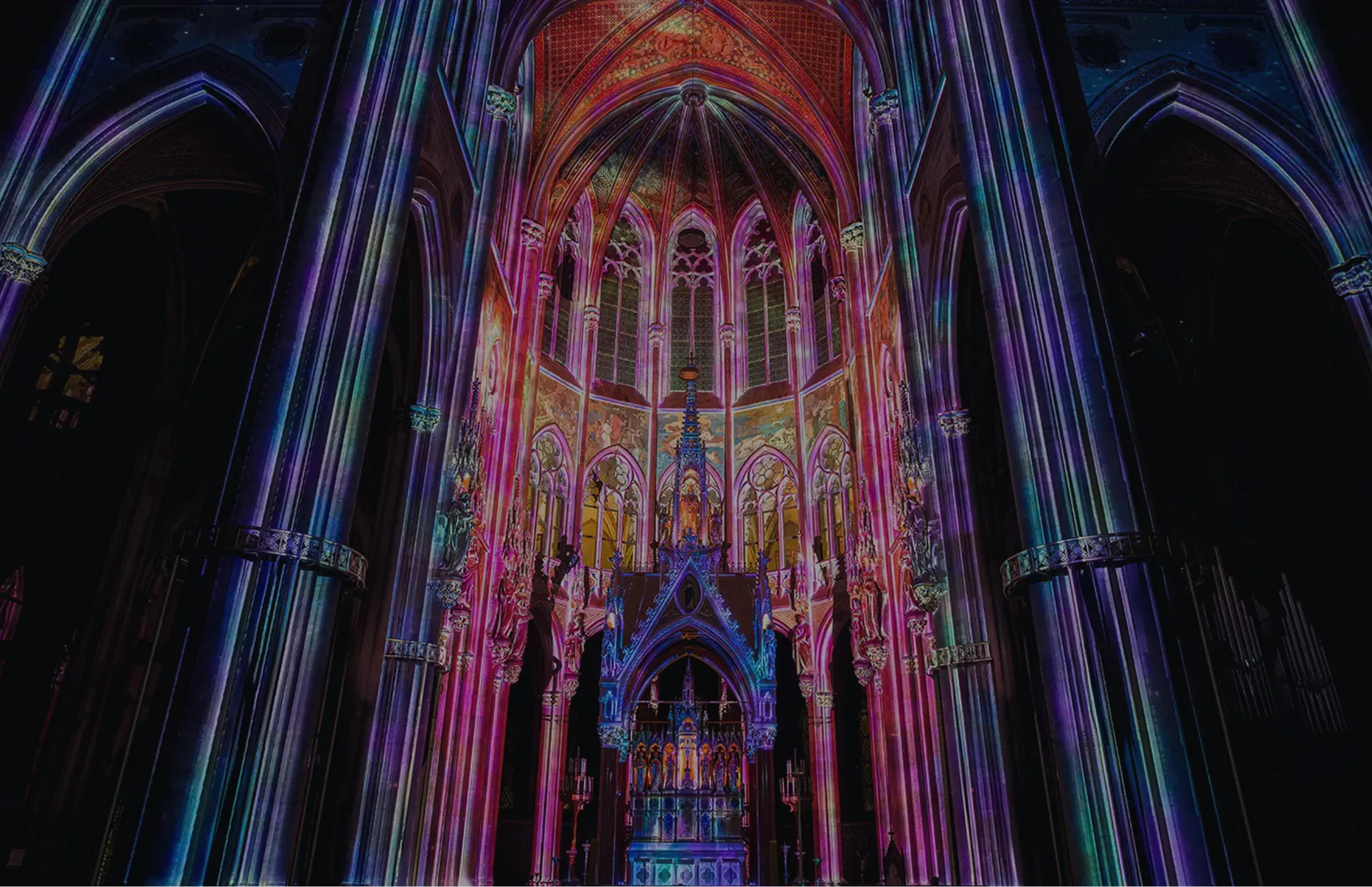 Vivid multicolor light projection illuminating the interior of a Gothic cathedral, with purple, pink, and blue hues highlighting the tall arches, stained glass, and ornate altar.