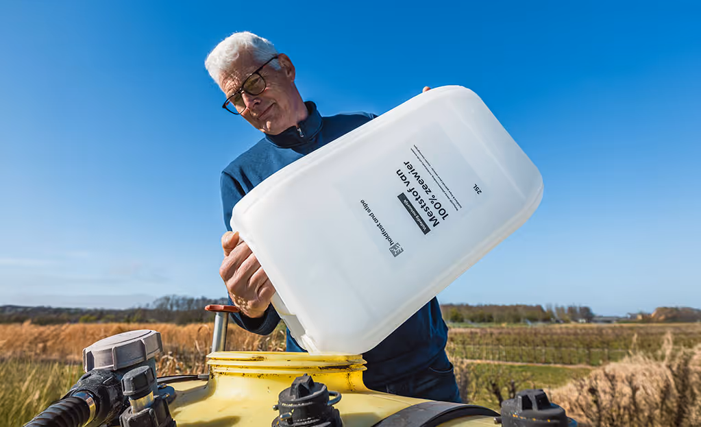 Een man met een bril giet 25 liter Holdfast and Stipe meststof in een gele landmachine op een veld.
