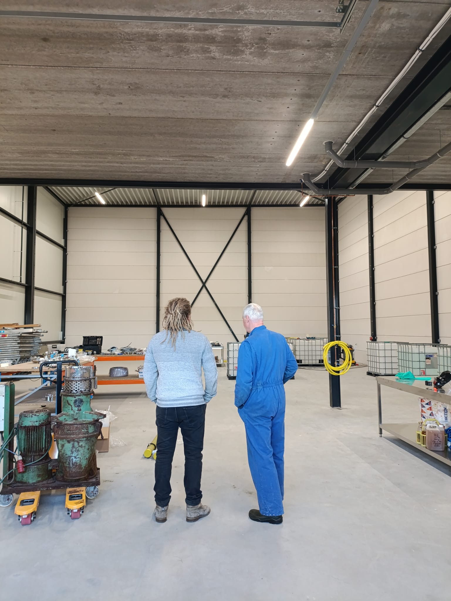 Twee mannen in een grote werkplaats met hoge plafonds, een man draagt een blauwe overall en de andere een grijze trui en zwarte broek.