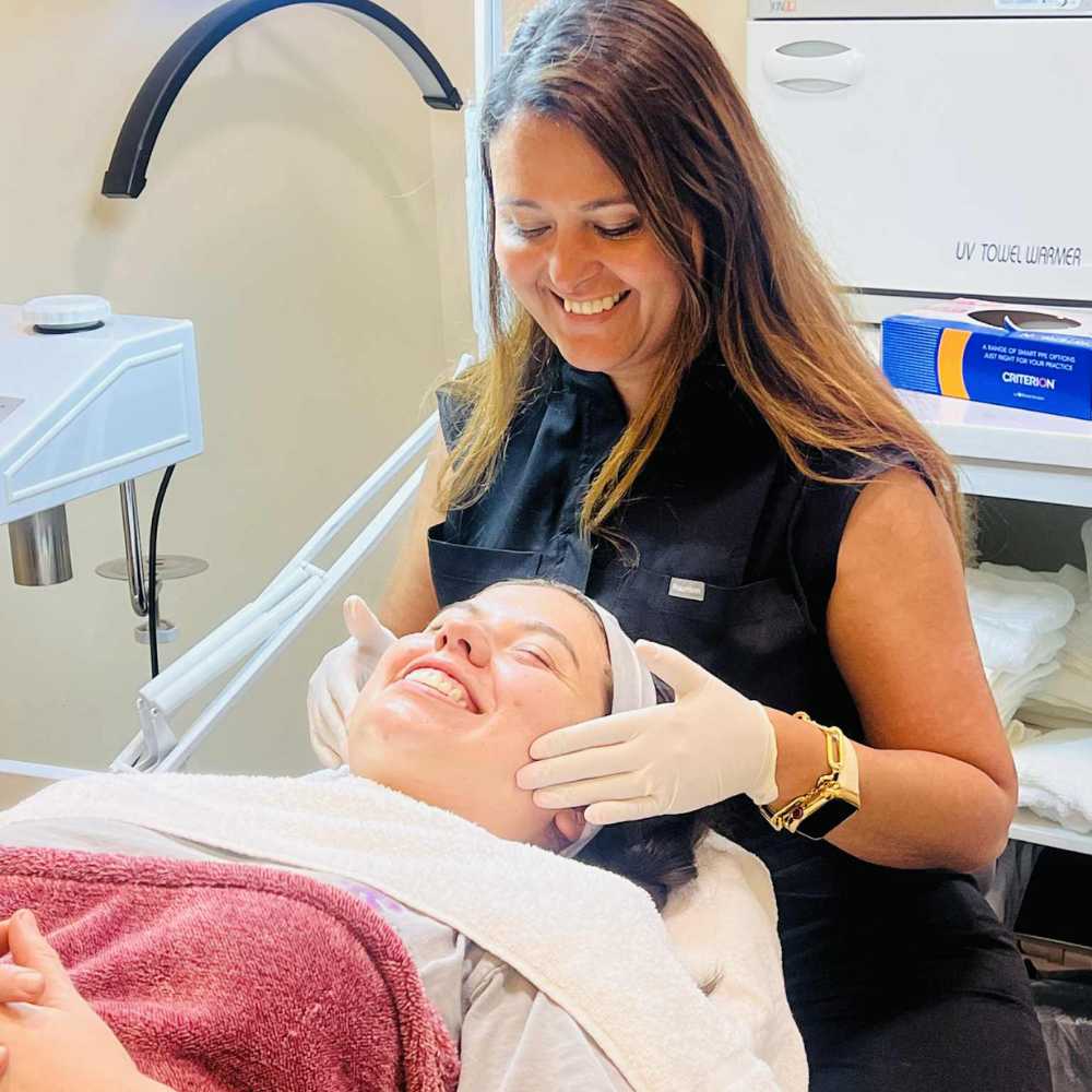 Smiling esthetician wearing gloves gently massages the face of a relaxed client lying on a treatment bed during a facial session.