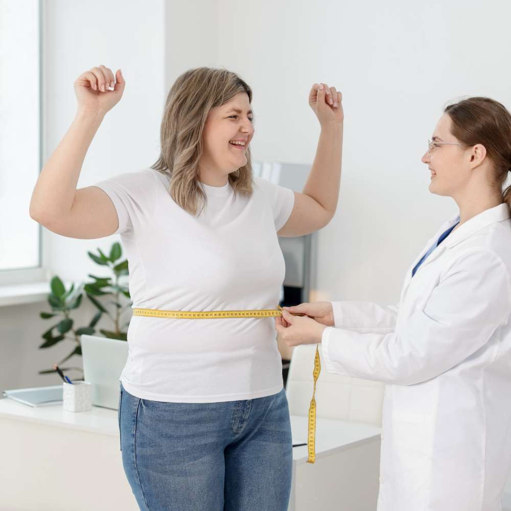 Smiling woman with arms raised while doctor measures her waist with a tape measure in a bright office at fanous medspa.