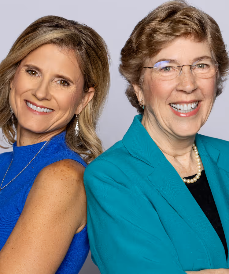 Two smiling women, one with blonde hair wearing a blue sleeveless top and the other with short light brown hair wearing glasses, a teal blazer, and a pearl necklace.