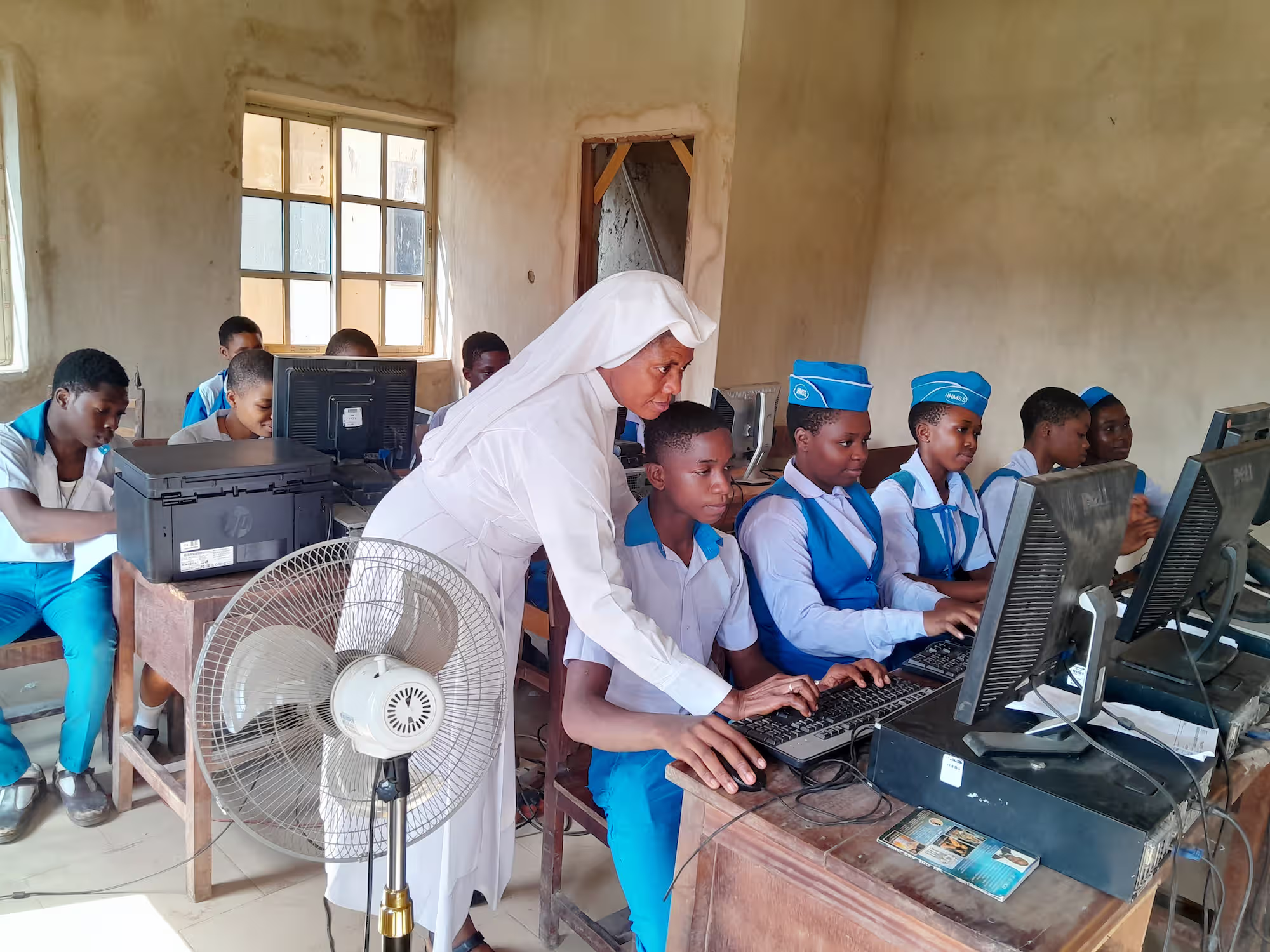 A nun in white assisting students in blue and white uniforms working on computers in a classroom.