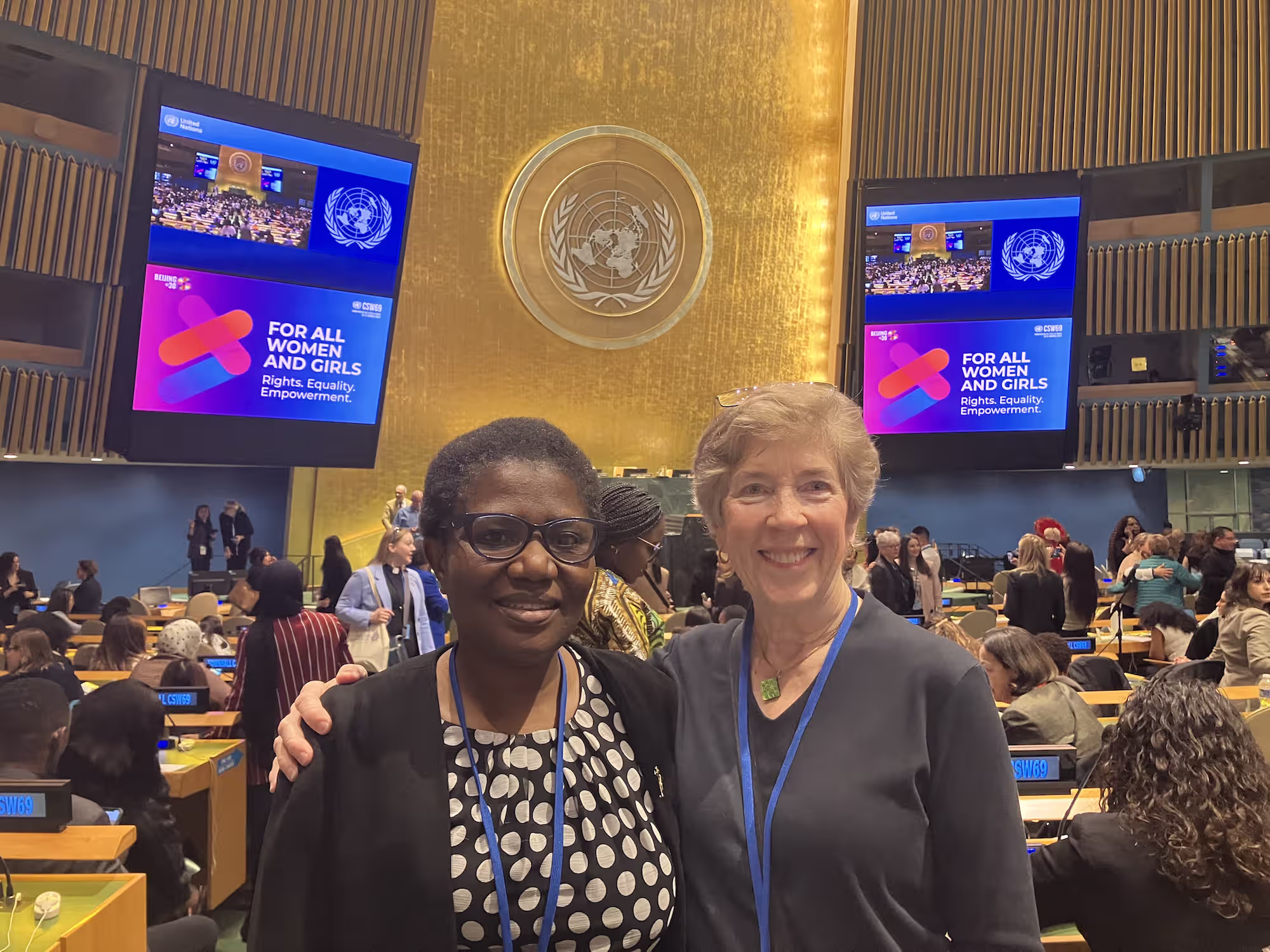 Two women smiling and posing with arms around each other in a conference hall with United Nations emblem and screens displaying 'For All Women and Girls. Rights. Equality. Empowerment.'