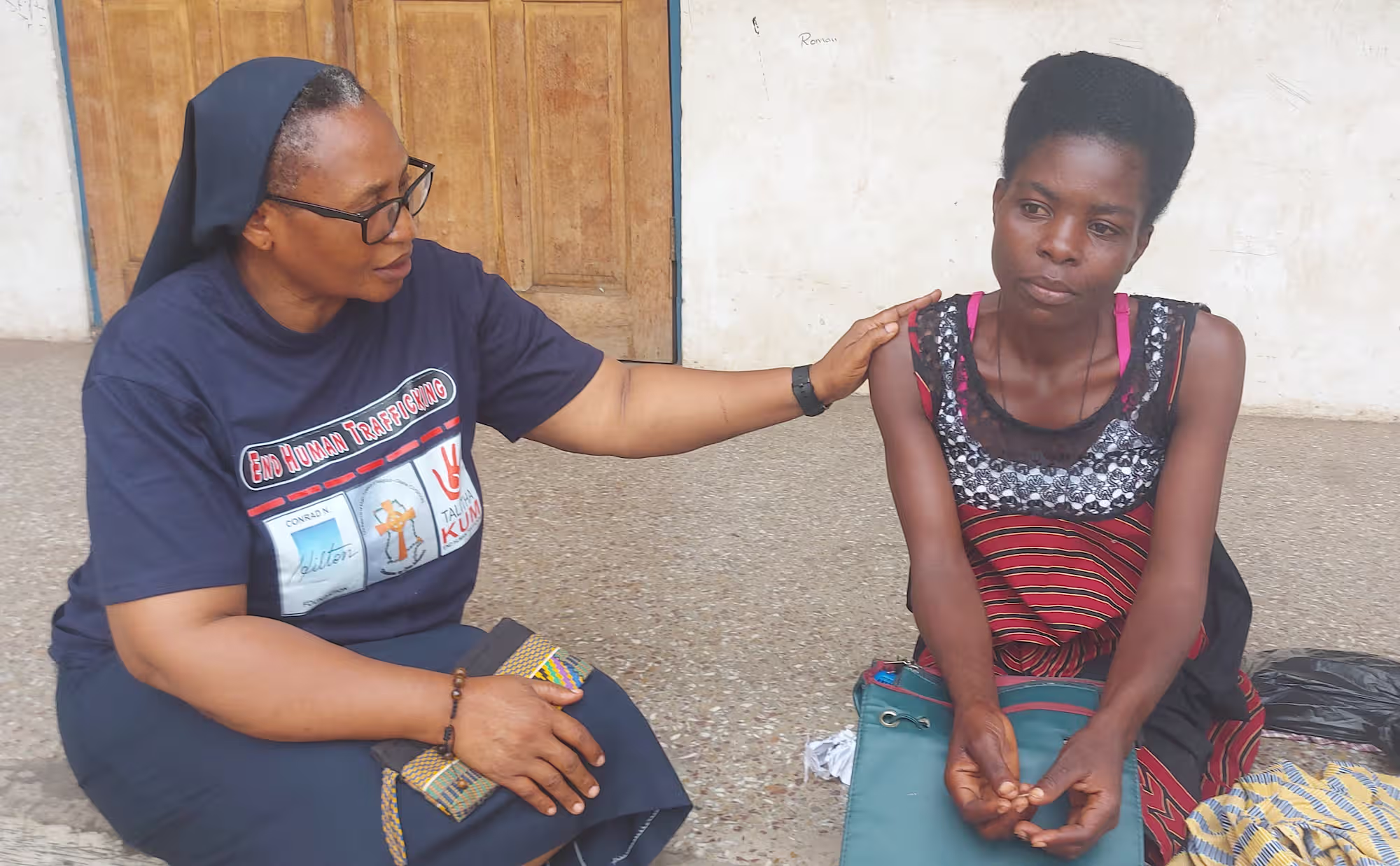 A woman wearing a navy t-shirt that reads 'End Human Trafficking' comforts a seated young woman with a serious expression.