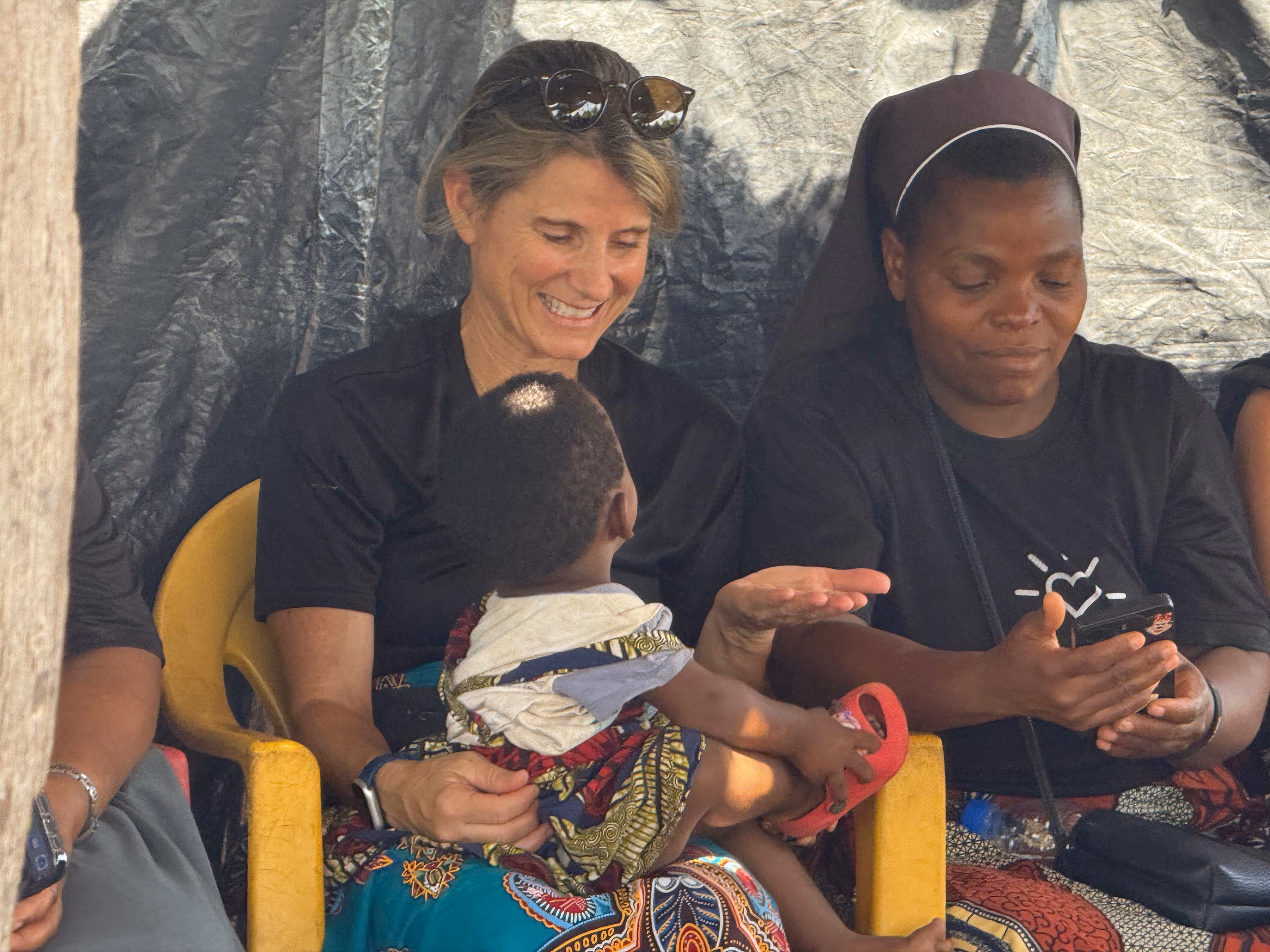 A woman smiling and holding a child on her lap while a nun beside her looks at a phone.