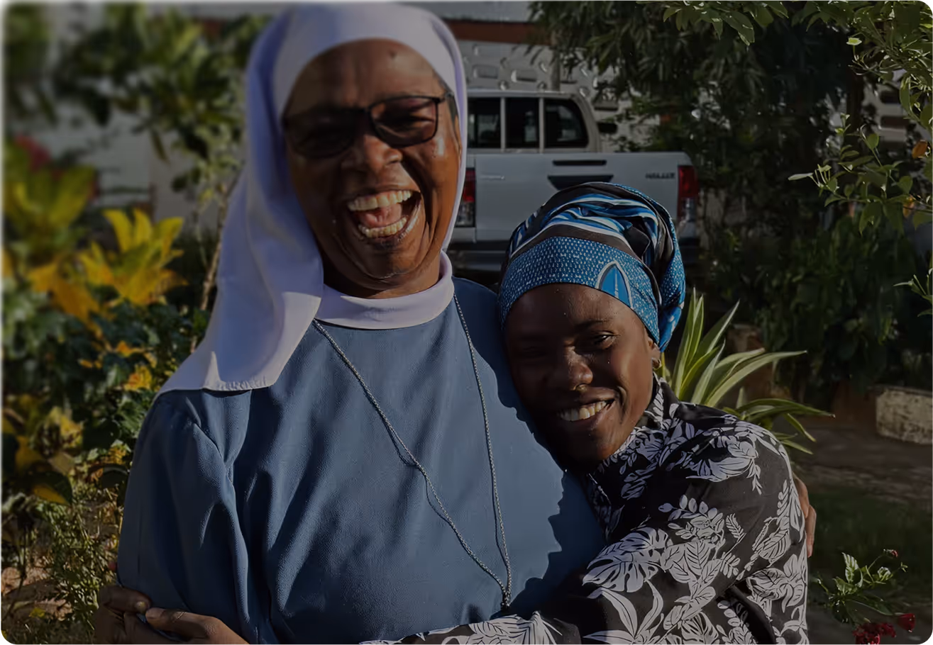 Two women smiling and hugging outdoors, one wearing a nun's habit and glasses, the other wearing a blue patterned headscarf and floral shirt.