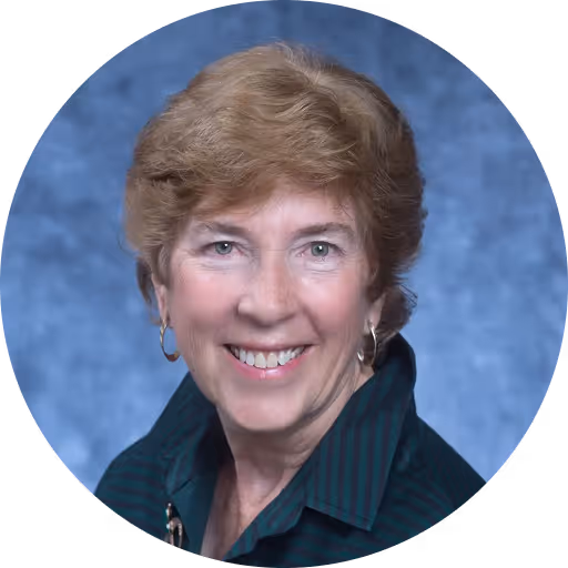 Smiling middle-aged woman with short light brown hair wearing hoop earrings and a dark green shirt against a blue background.