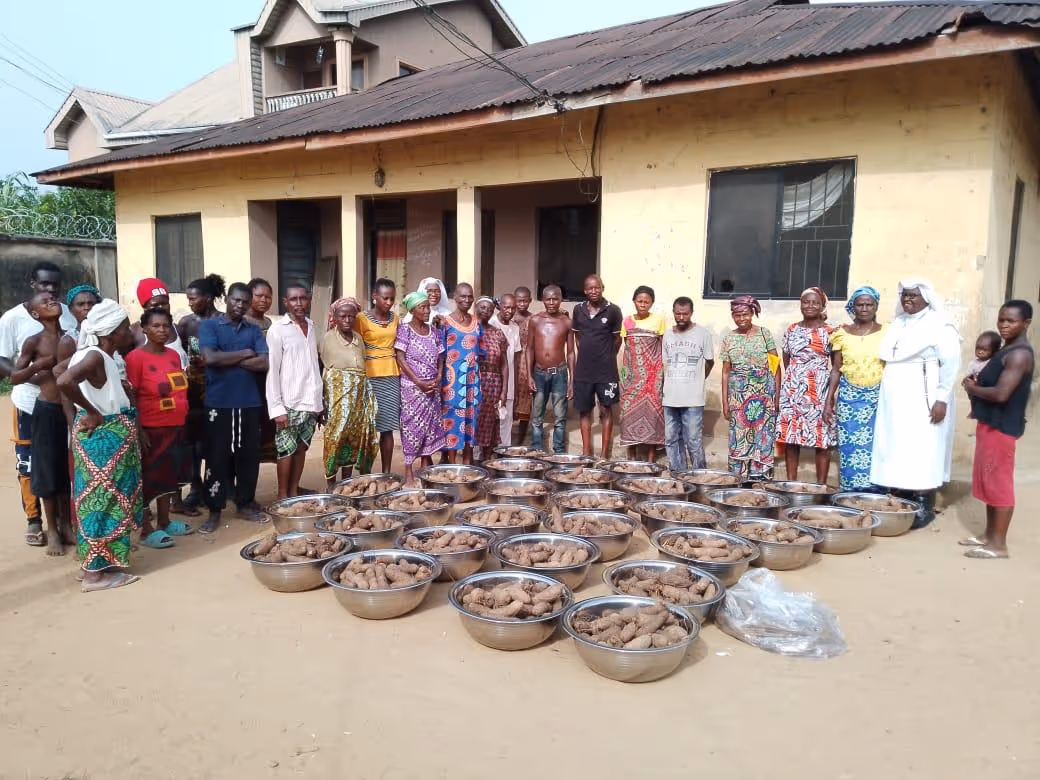 Group of people standing in front of a building with multiple large metal bowls filled with harvested yams on the ground.