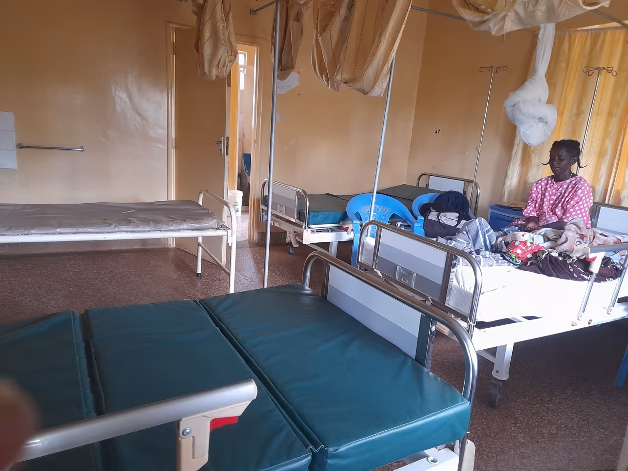 Hospital room with multiple beds and a woman sitting on a bed covered with blankets.