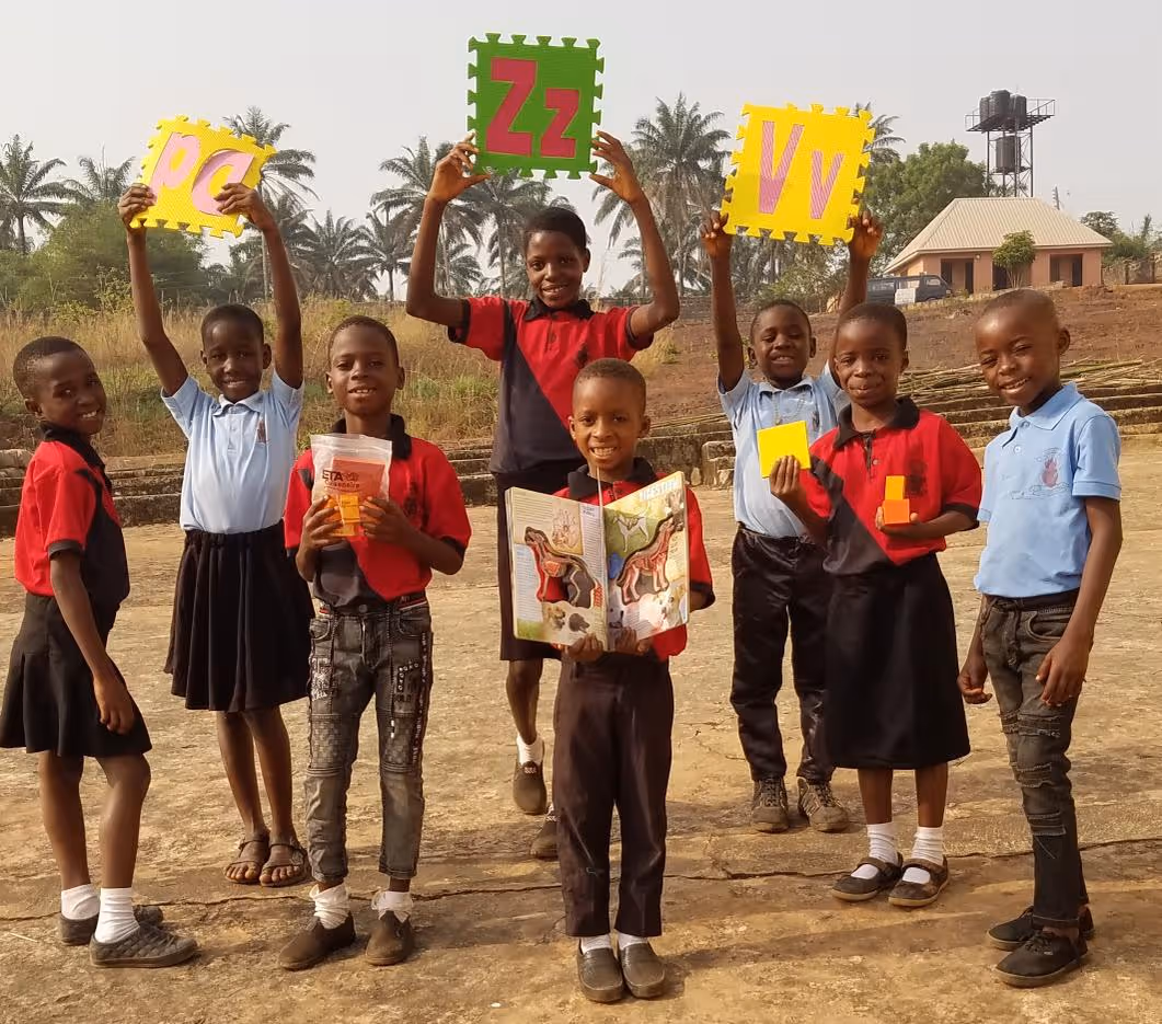 Eight smiling children standing outdoors holding educational materials including alphabet puzzle pieces and a picture book.