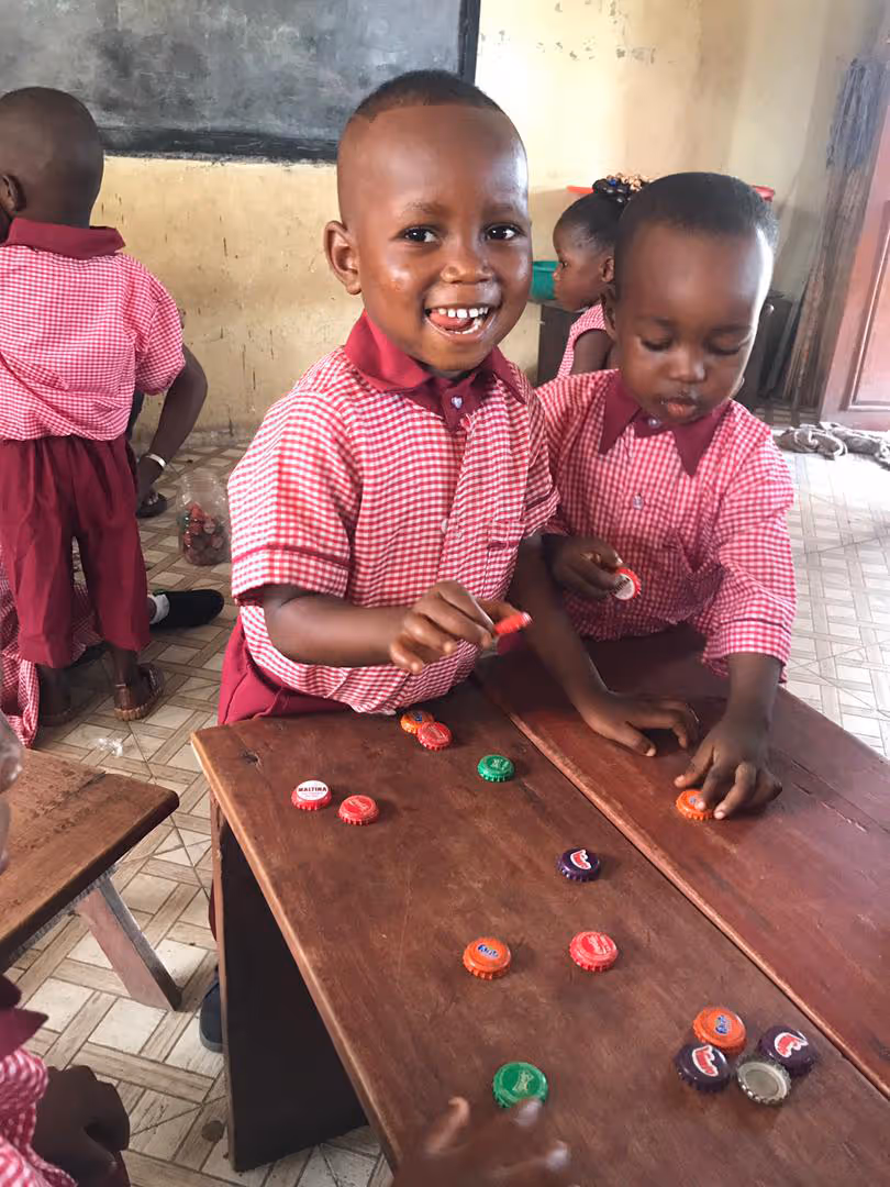 Young children in matching red checkered uniforms playing with colorful bottle caps on wooden desks in a classroom.