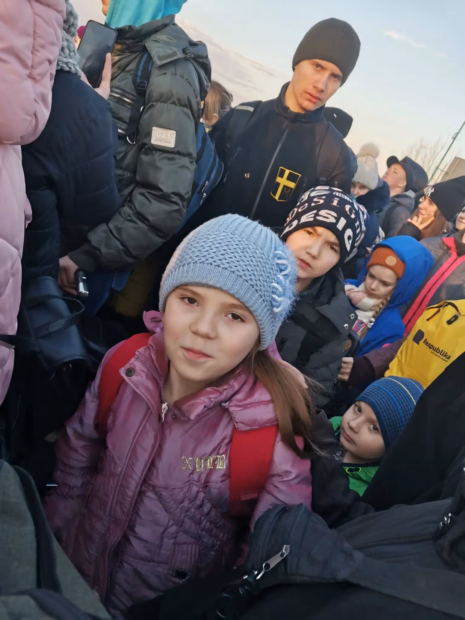 A crowd of people dressed in winter clothing, including children wearing hats and jackets, gathered closely together outdoors.