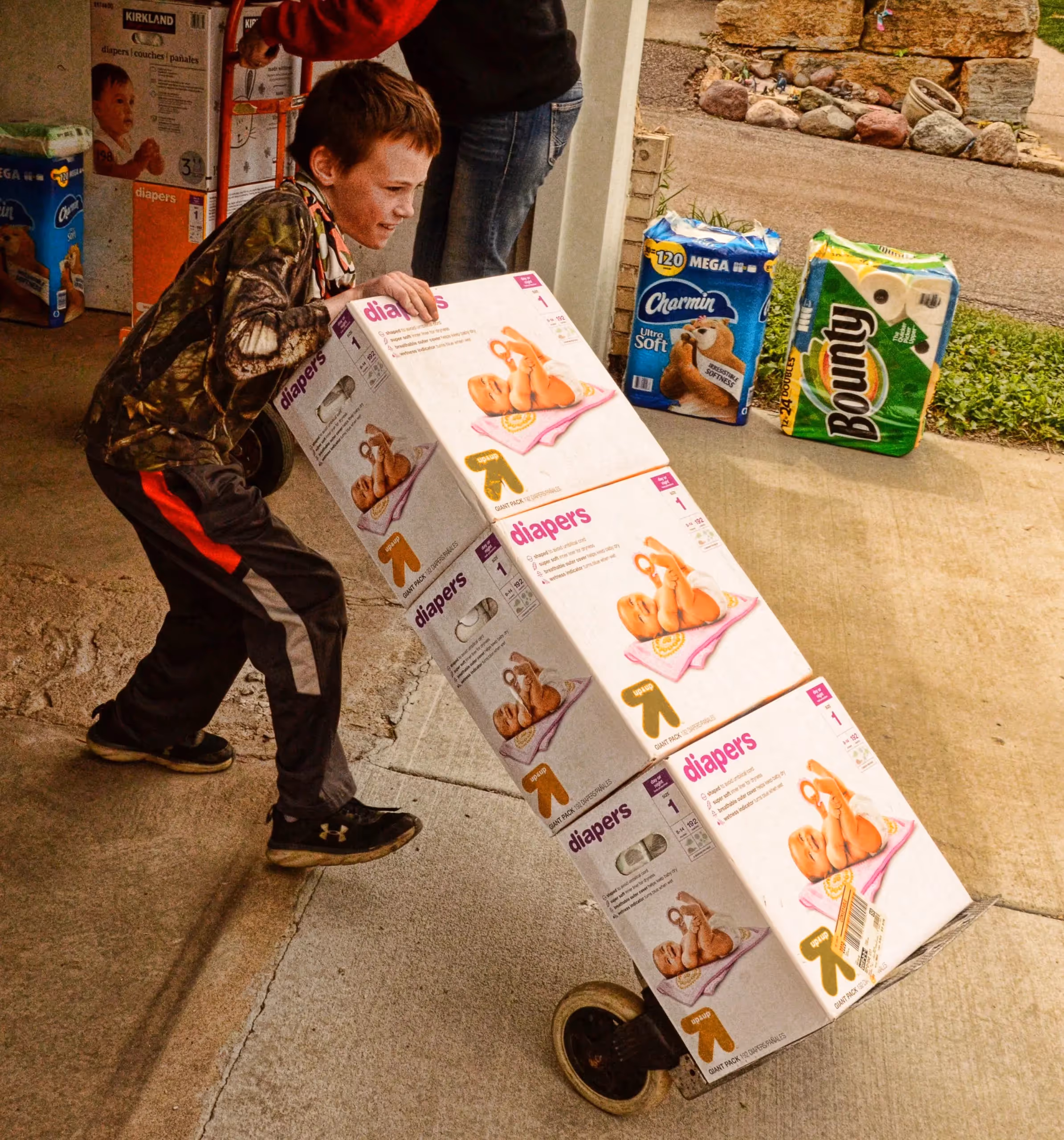 Young boy in camouflage shirt pushing a dolly stacked with large diaper boxes outside a building.