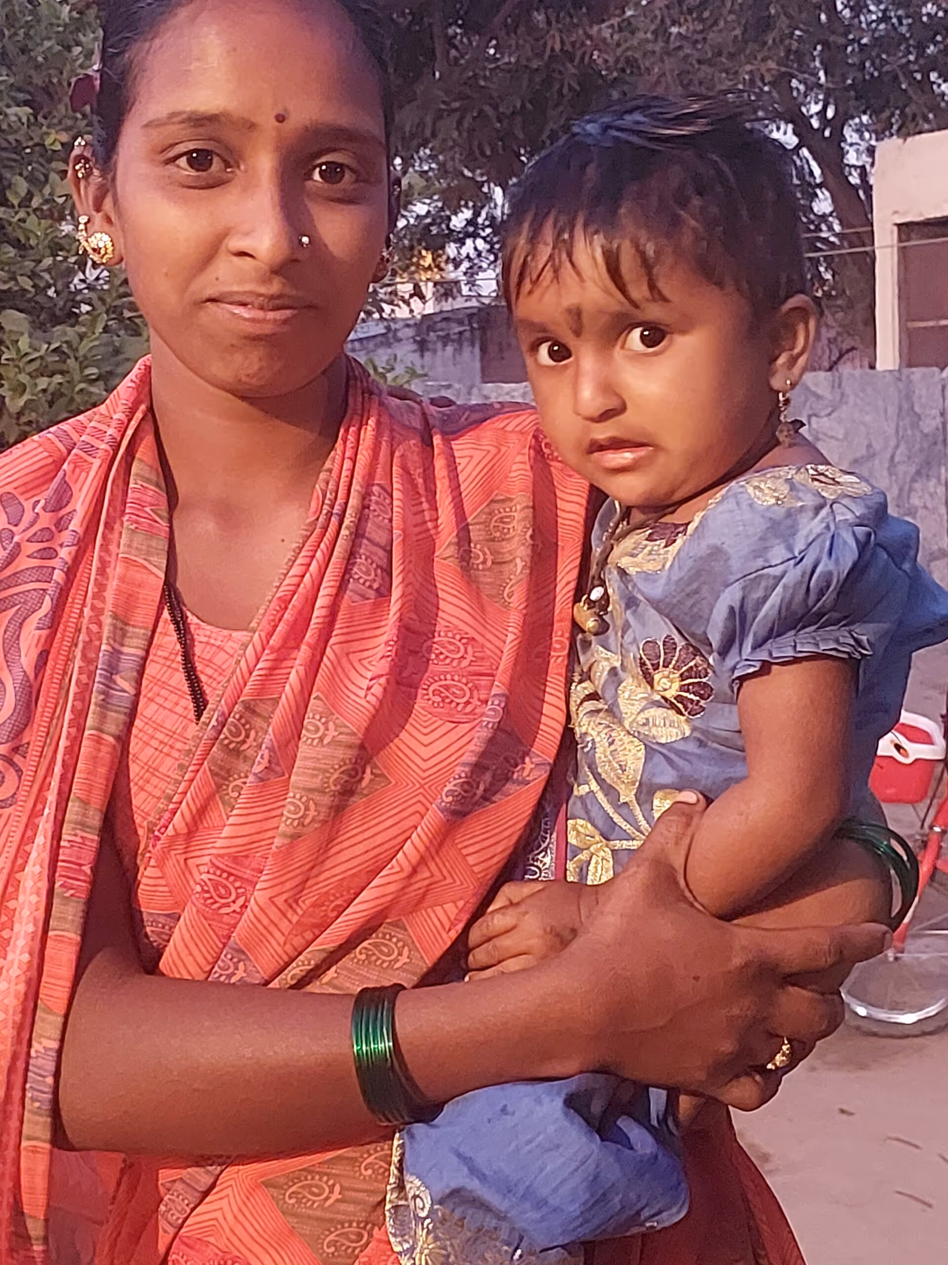 A woman in a patterned pink sari holding a toddler wearing a blue dress, both looking at the camera outdoors.