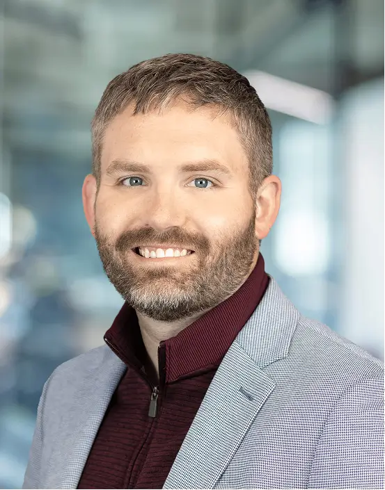 Smiling woman with shoulder-length light brown hair wearing a blue top and black cardigan in a softly blurred indoor setting.