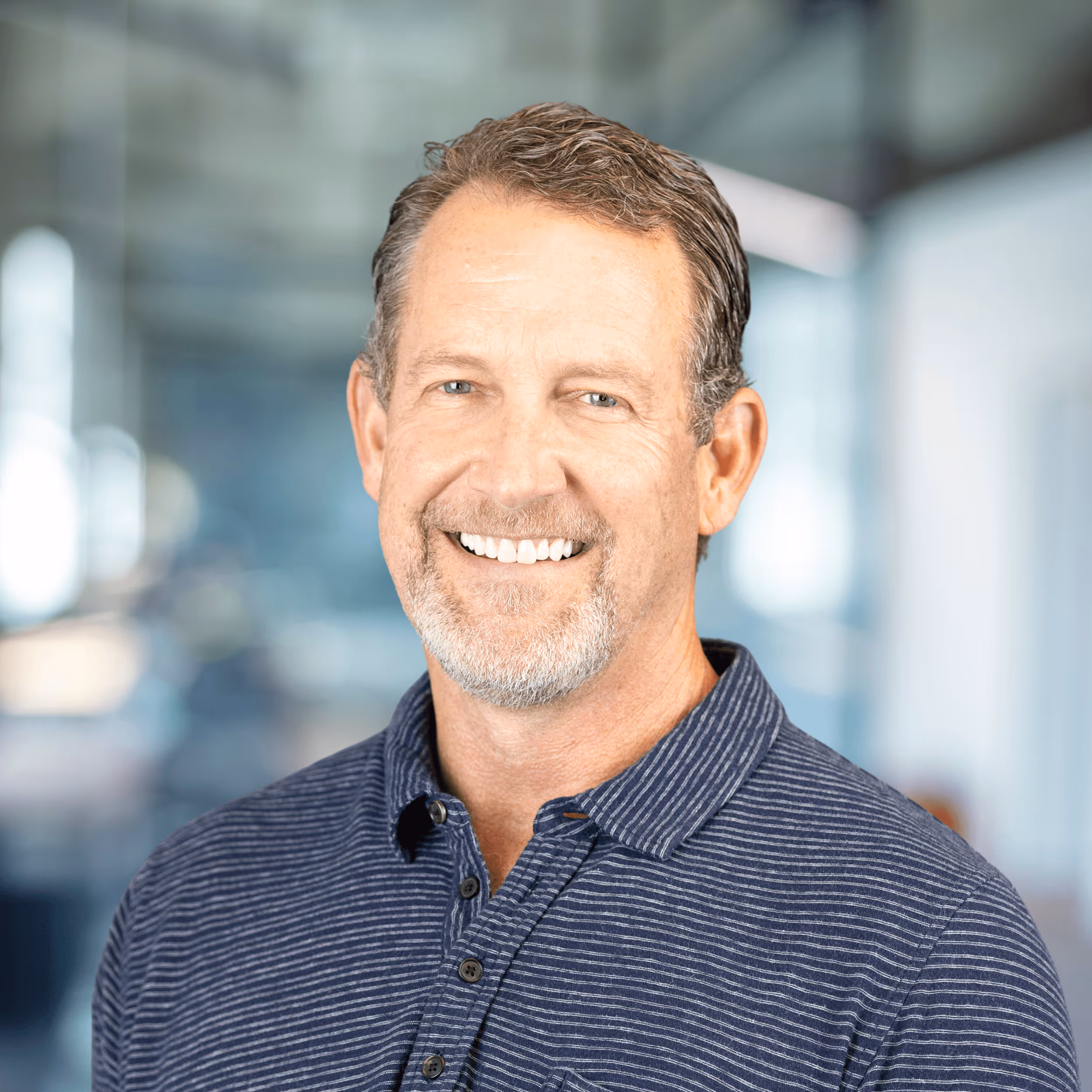 Smiling man with short brown hair and beard wearing a gray blazer over a maroon zip-up shirt, standing in a bright modern office.