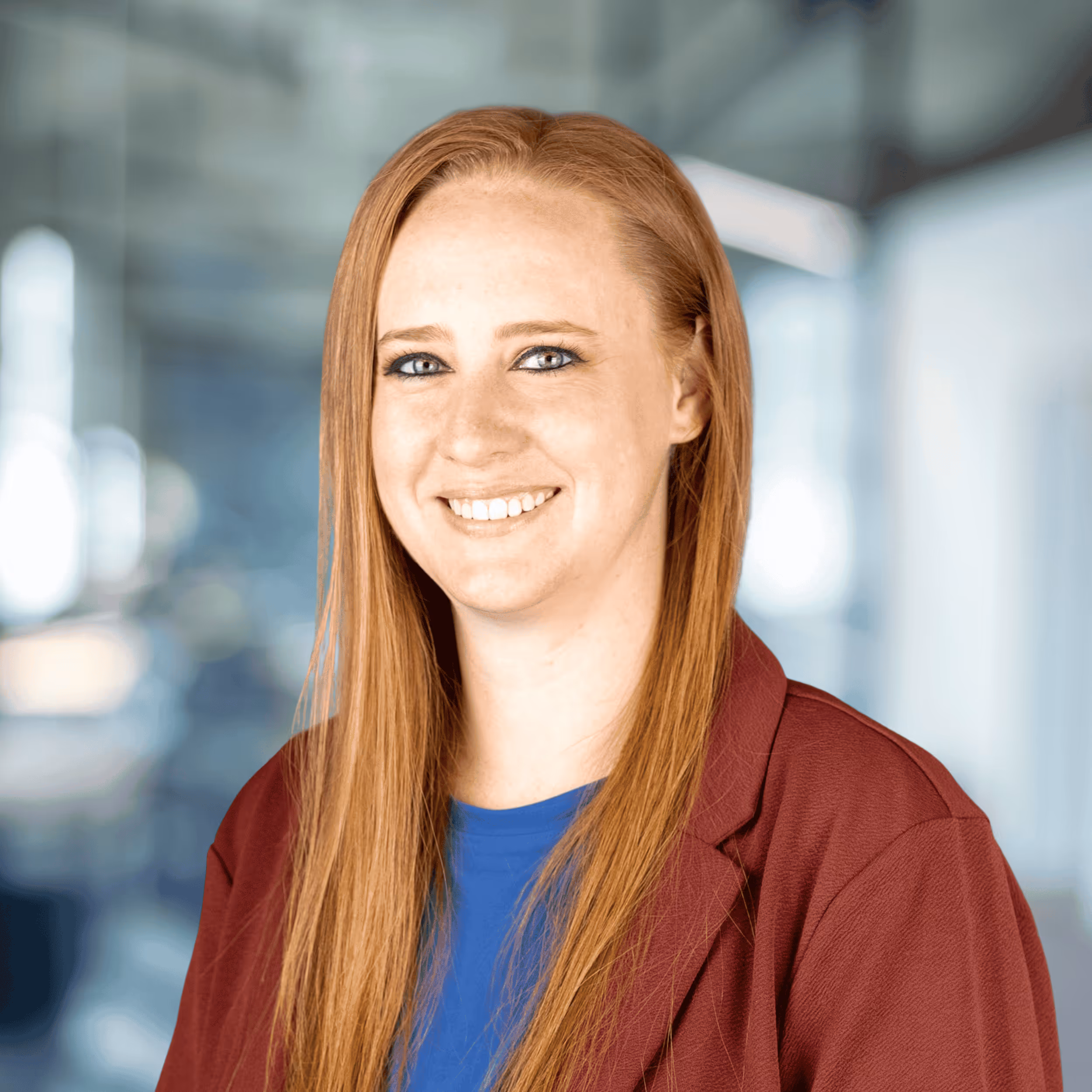 Smiling woman with long red hair wearing a maroon blazer and blue top in a modern office setting.