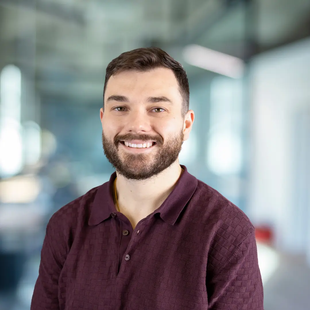 Smiling man with short brown hair and beard wearing a light gray jacket over a maroon zip-up top in a blurred office setting.