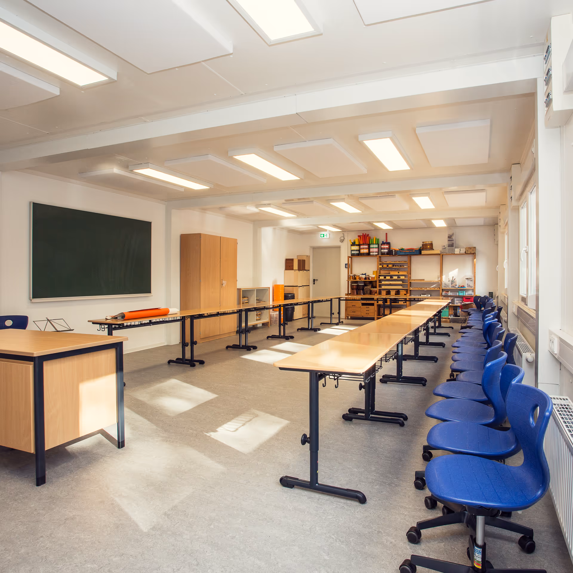 Heller Klassenraum mit grüner Tafel, langen Tischen und einer Reihe blauer Stühle auf Rollen.