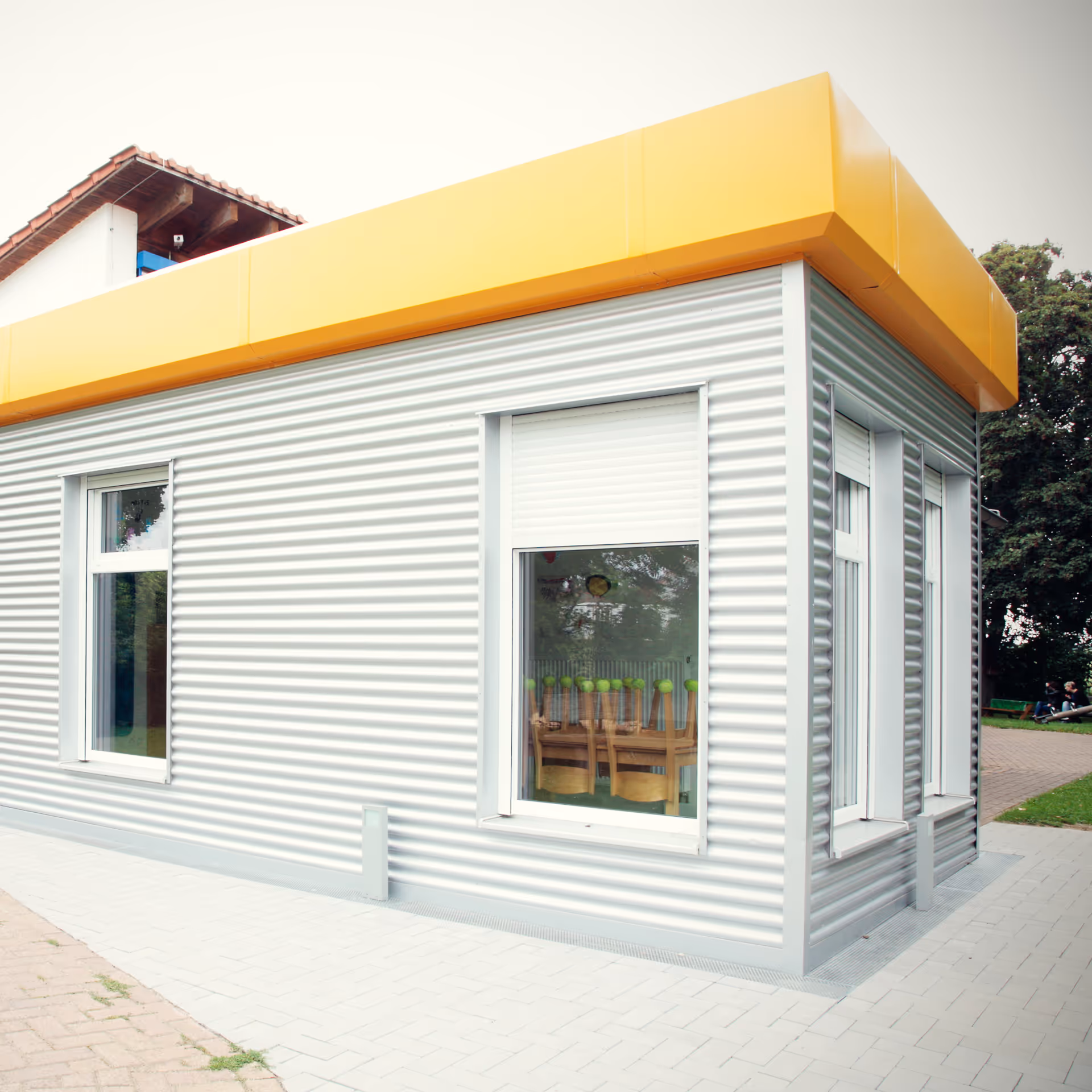 Eckansicht eines Gebäudes mit grauen, wellenförmigen Metallwänden, weißen Fenstern und gelbem Dachrand.