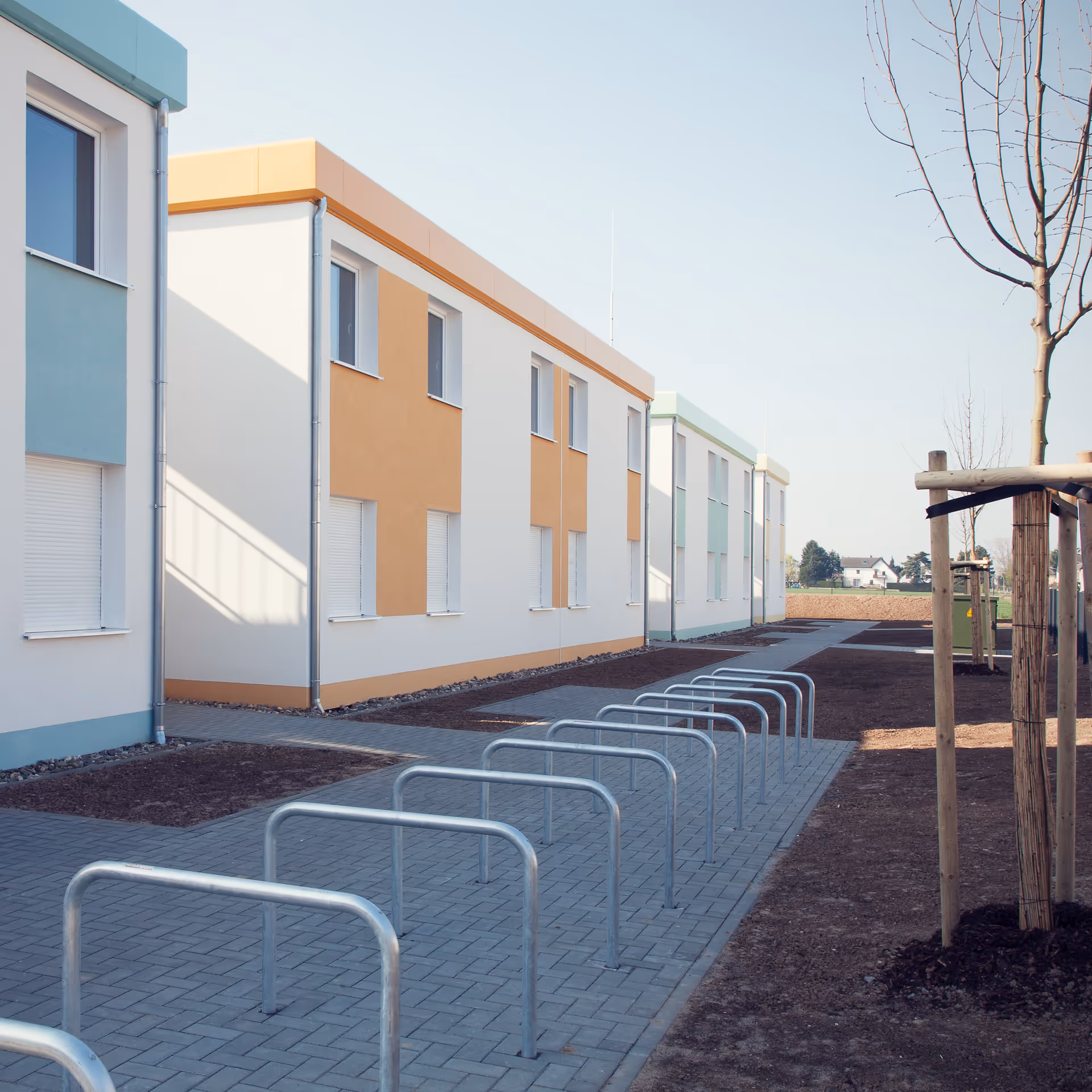 Reihe moderner, zweistöckiger Wohngebäude mit pastellgelb und pastellgrün akzentuierten Fassaden und Fahrradständern auf einem gepflasterten Weg.