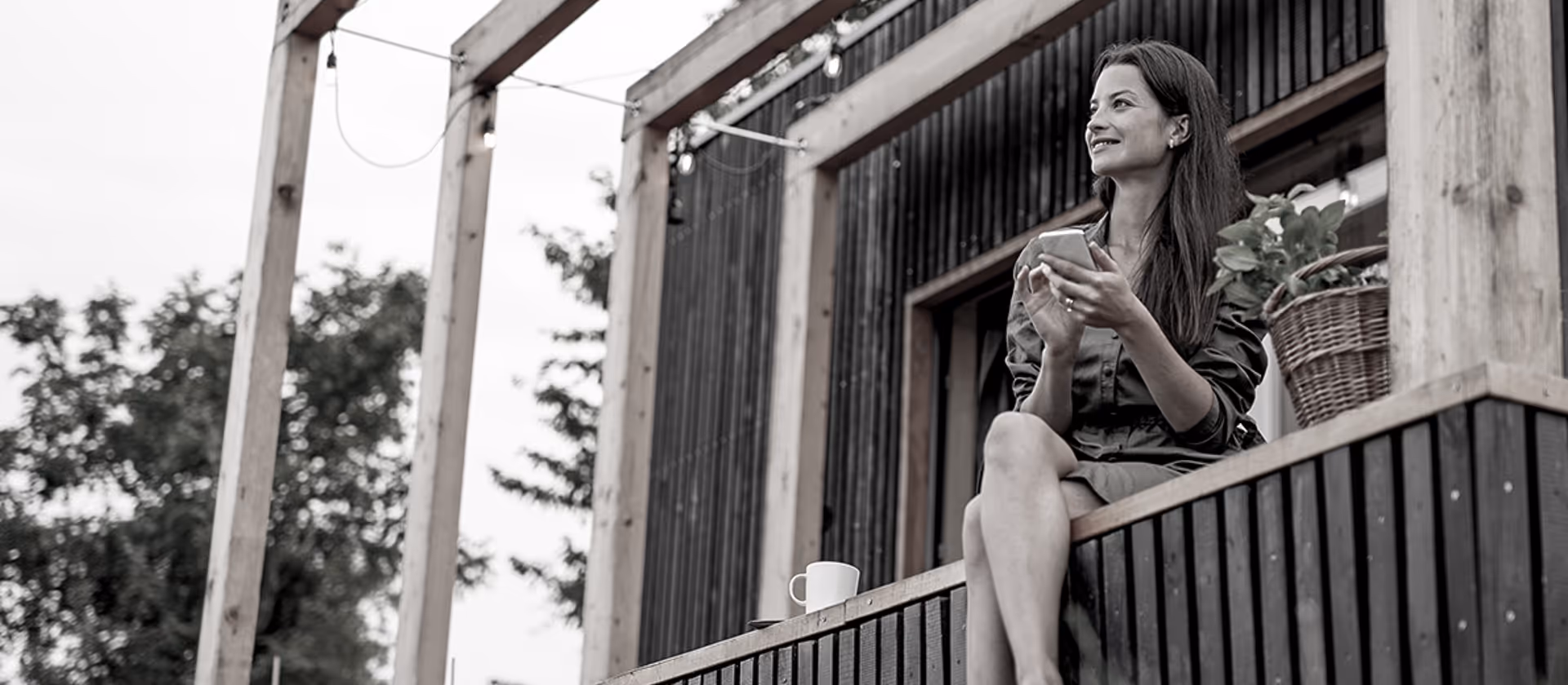 Frau sitzt auf einer Veranda mit einem Smartphone in der Hand und lächelt, mit einer Pflanze im Korb neben ihr.