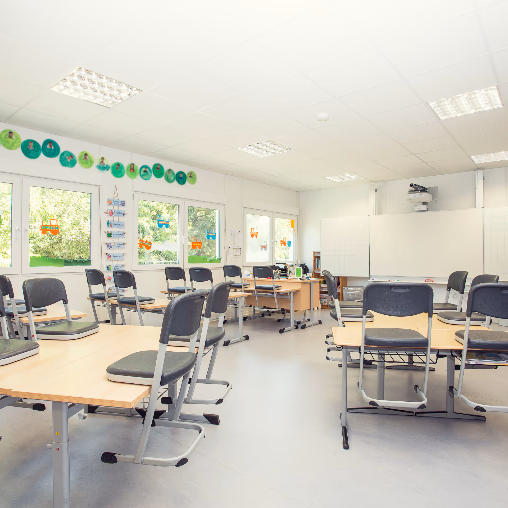 Helle Schulklasse mit Stühlen über Tischen, weißen Wänden, großen Fenstern mit Kindermalereien und einem Whiteboard mit Projektor.
