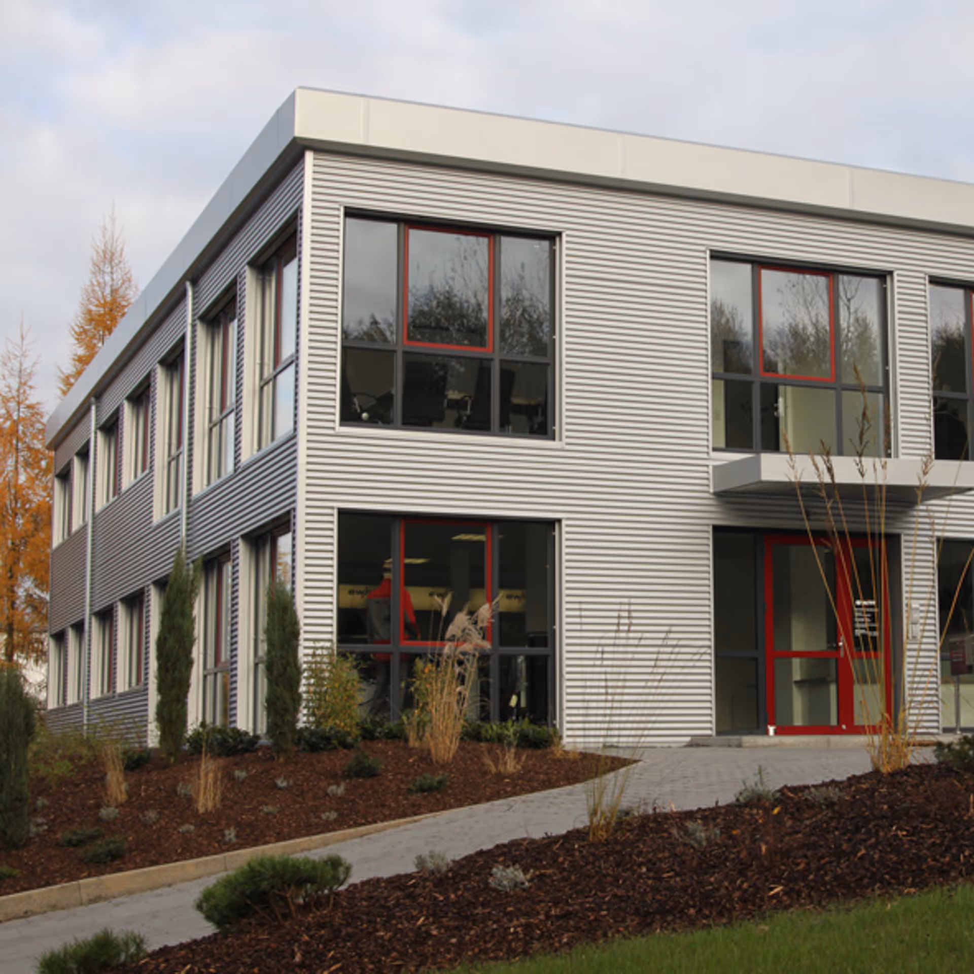 Modernes zweistöckiges Bürogebäude mit grauer Metallfassade, roten Fensterrahmen und einem gepflasterten Gehweg davor.