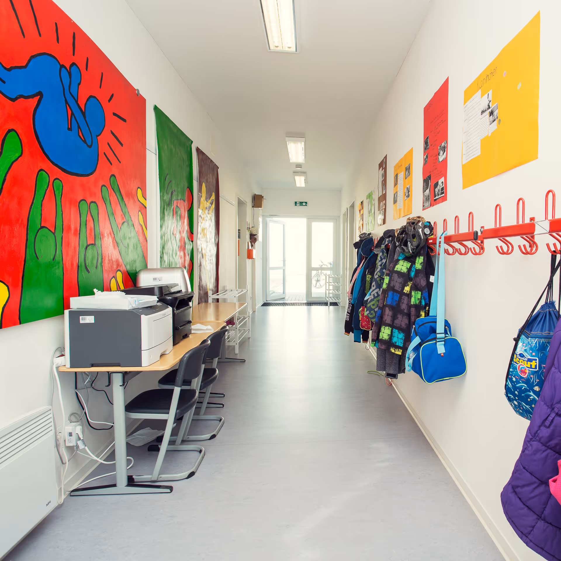 Flur mit Kinderjacken, Taschen an roten Haken, bunte Wandbilder und Tische mit Druckern in einer hellen Schule.