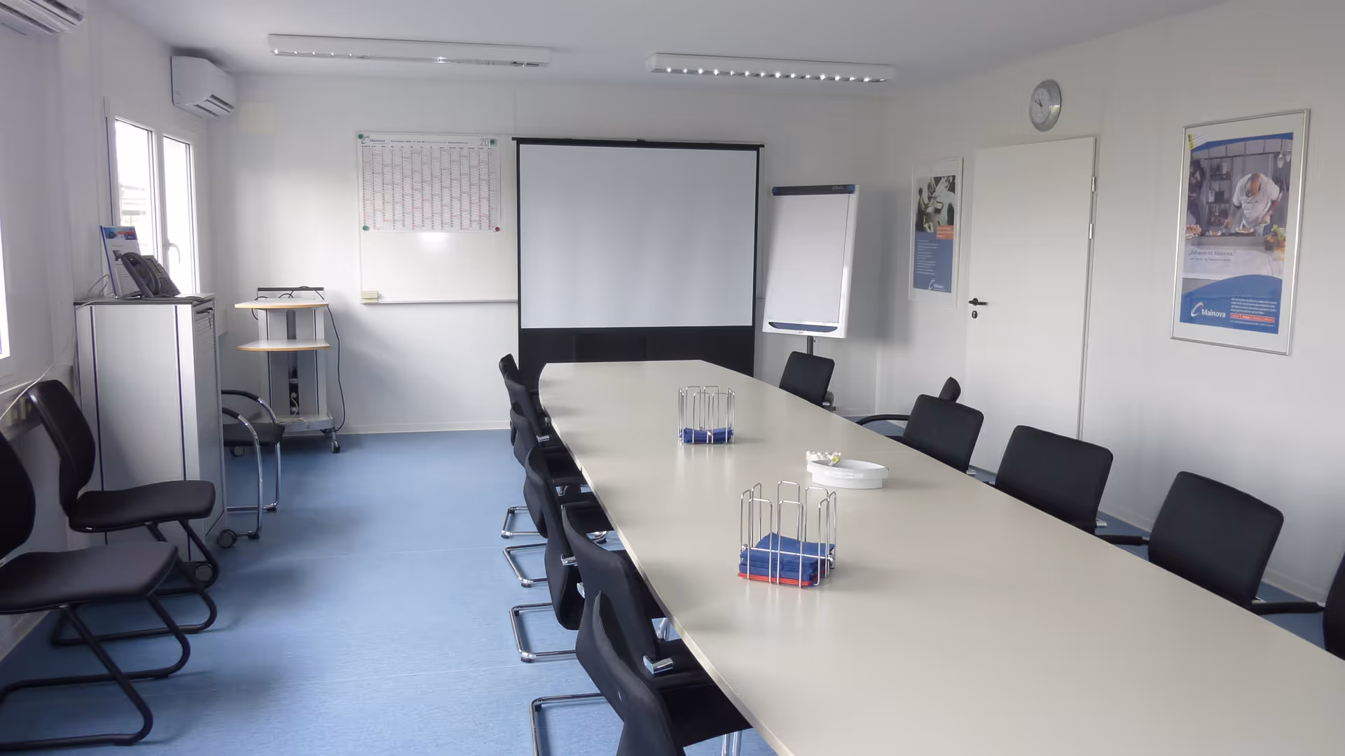 Leerer Konferenzraum mit langem weißen Tisch, schwarzen Stühlen, Whiteboard, Flipchart und Projektionsleinwand.