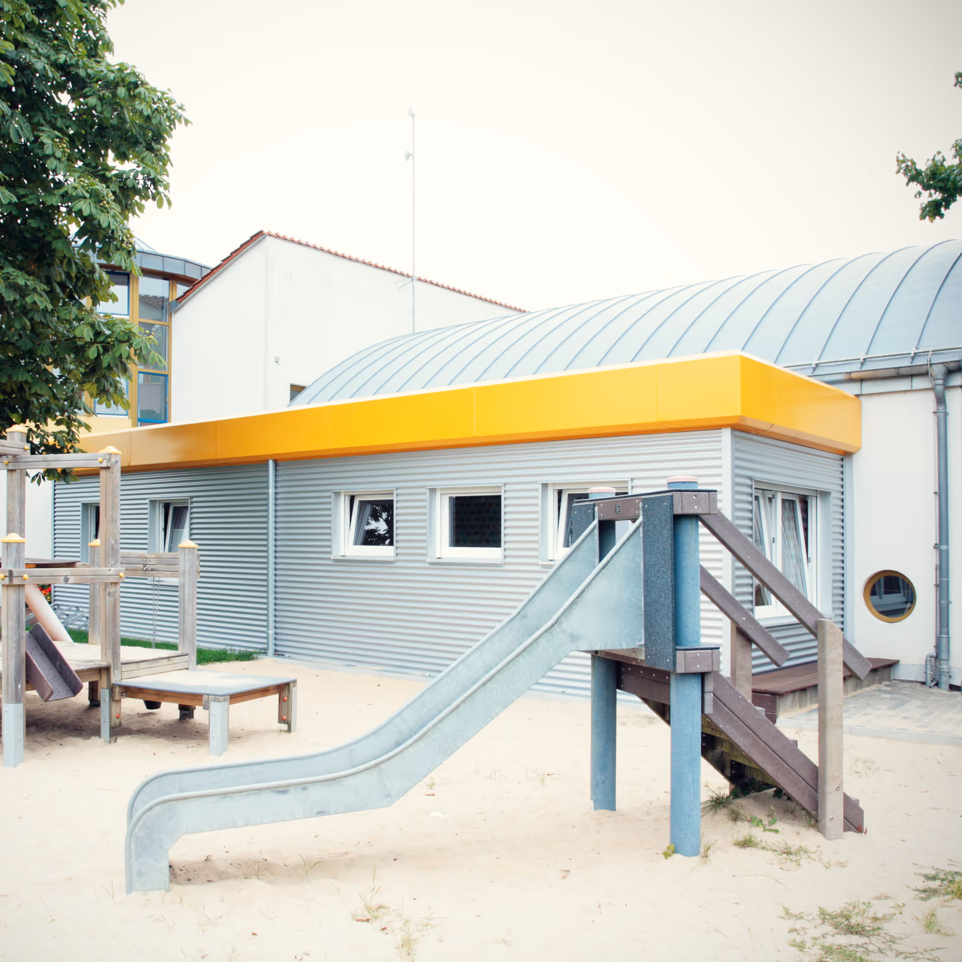 Kinderspielplatz mit einer Metallrutsche und einem Klettergerüst vor einem Gebäude mit grauer Fassade und gelbem Dachrand.