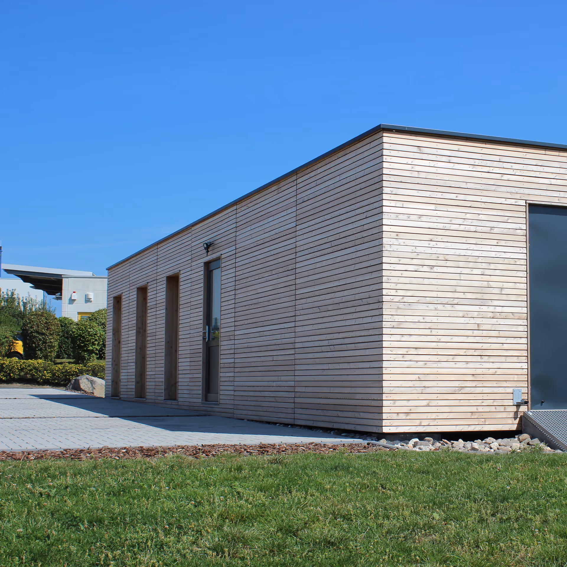 Modernes rechteckiges Gebäude mit Holzverkleidung, Türen und einem grasbewachsenen Vordergrund unter blauem Himmel.