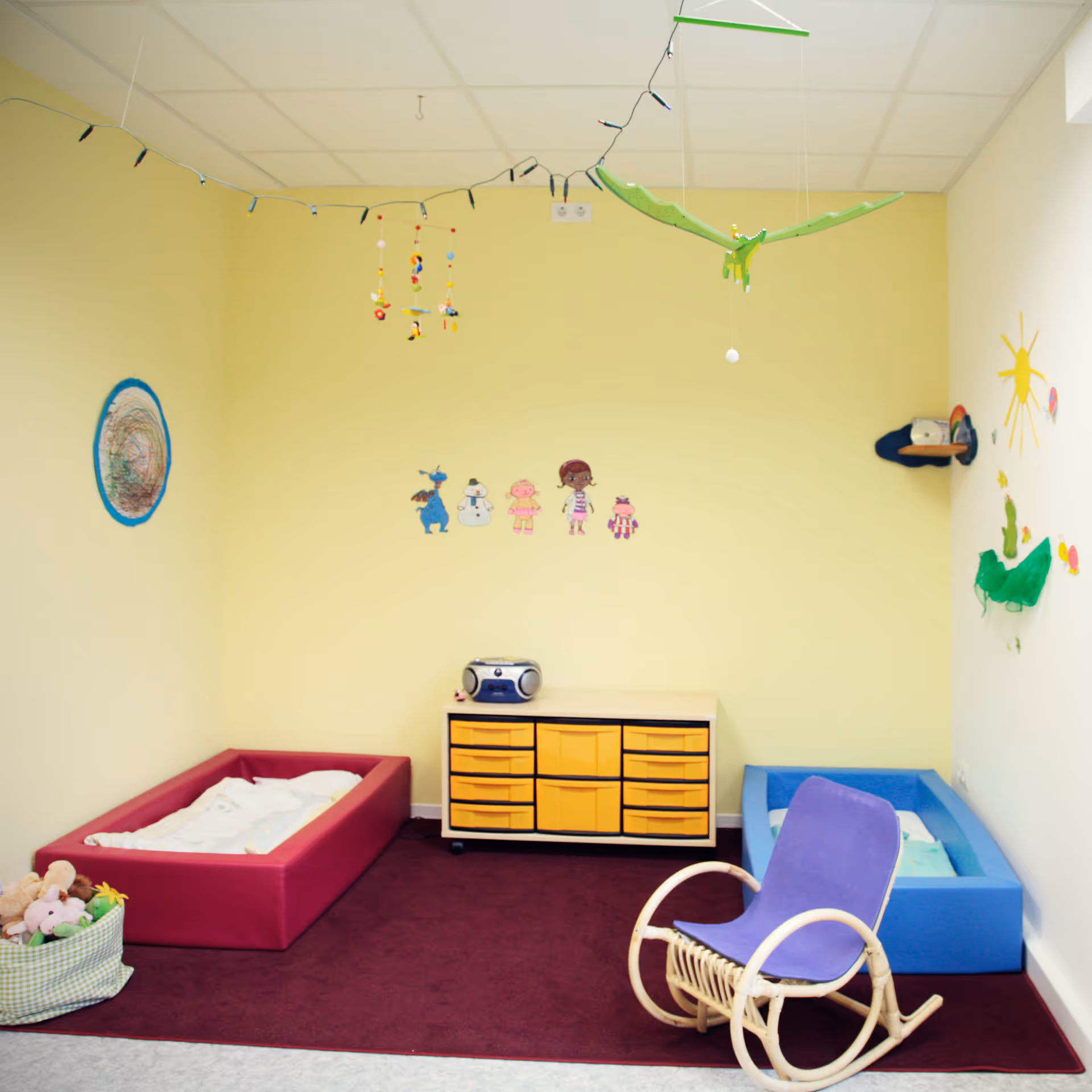 Bunter Kindergartenraum mit zwei kleinen Betten, einem violetten Schaukelstuhl, gelber Kommode und Wanddekorationen.