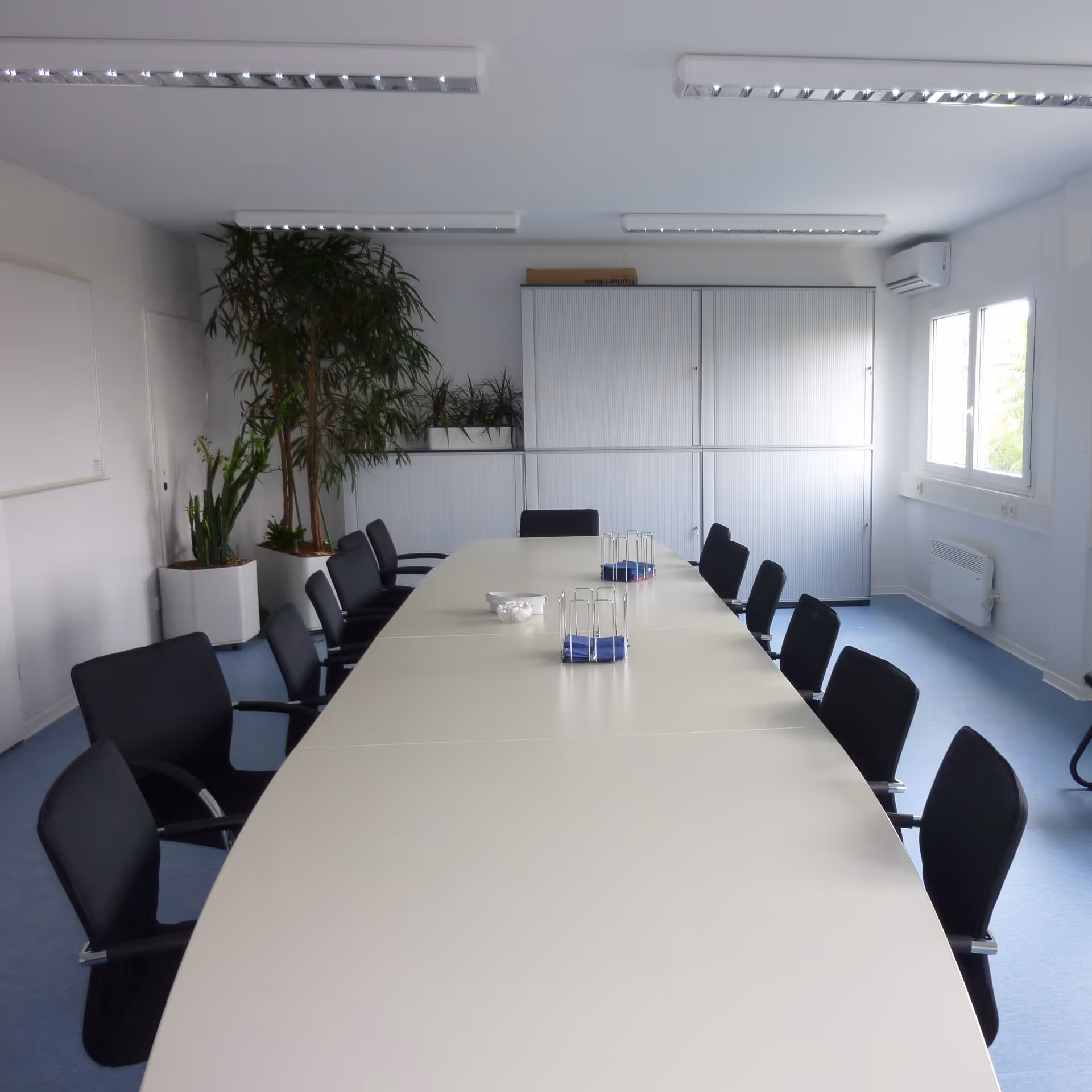 Moderner, leerer Konferenzraum mit langem weißen Tisch, schwarzen Stühlen und großen Pflanzen in den Ecken.