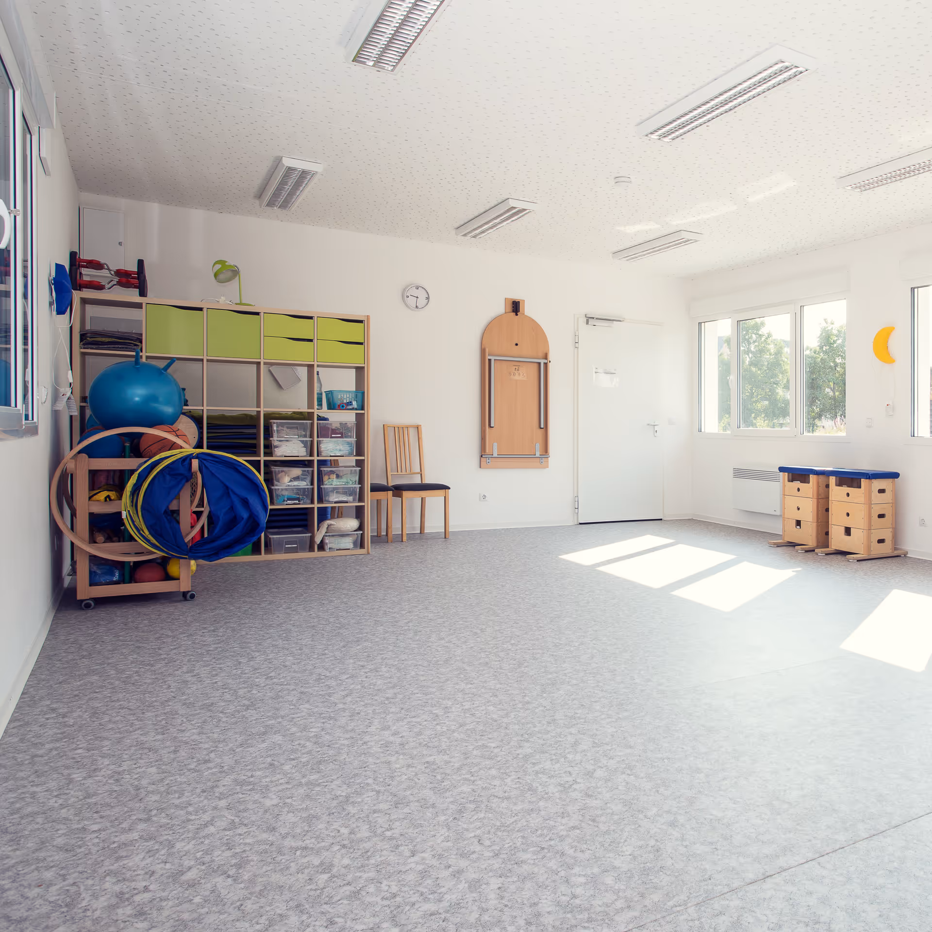 Heller, leerer Raum mit Sport- und Therapieausrüstung, Stuhl und großen Fenstern mit Blick auf Bäume.