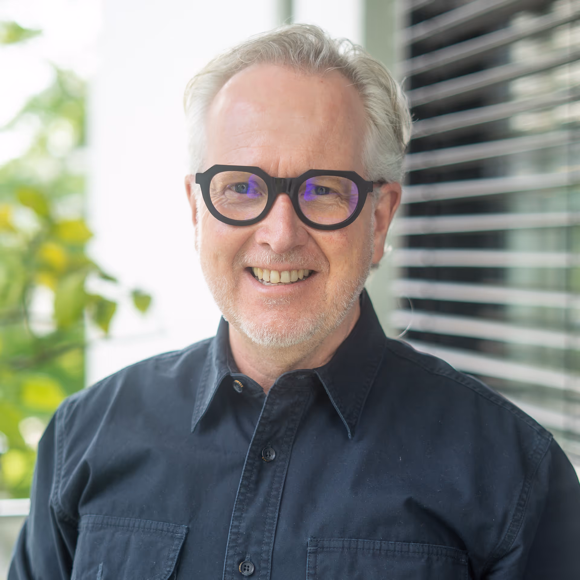 Älterer Mann mit weißen Haaren, Bart und runden schwarzen Brillengestellen, der lächelnd vor einem Fenster mit Jalousien und grünen Pflanzen im Hintergrund steht.