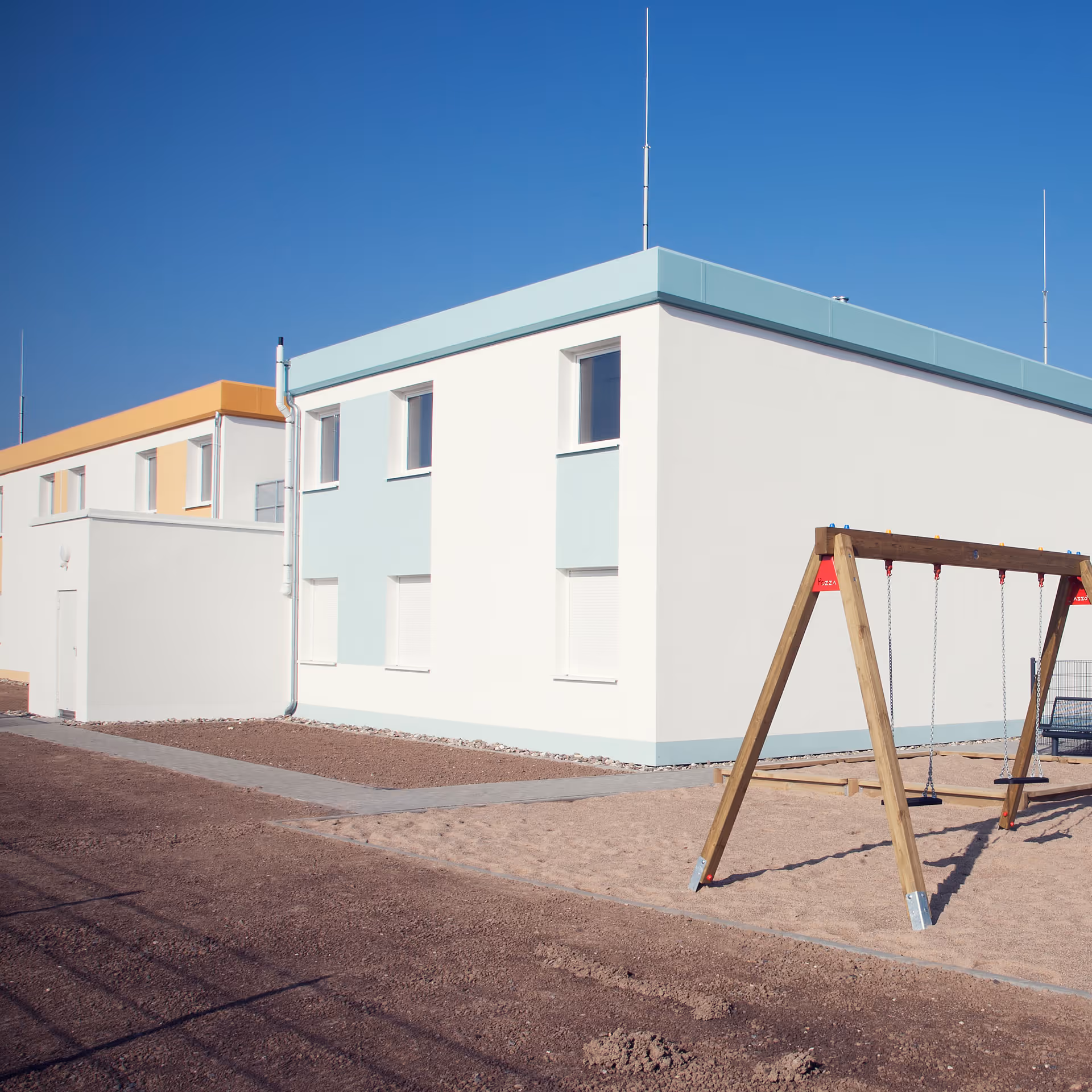 Ein moderner zweistöckiger Wohnblock mit weißer Fassade und blauen sowie orangefarbenen Akzenten, daneben eine Schaukel auf einem Sandspielplatz.