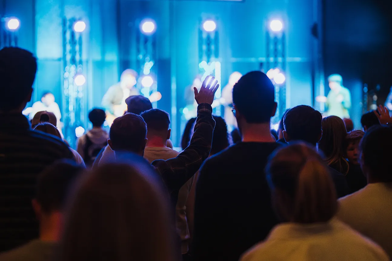 Audience at a concert or event with one person raising their hand, bright stage lights and performers blurred in blue light.