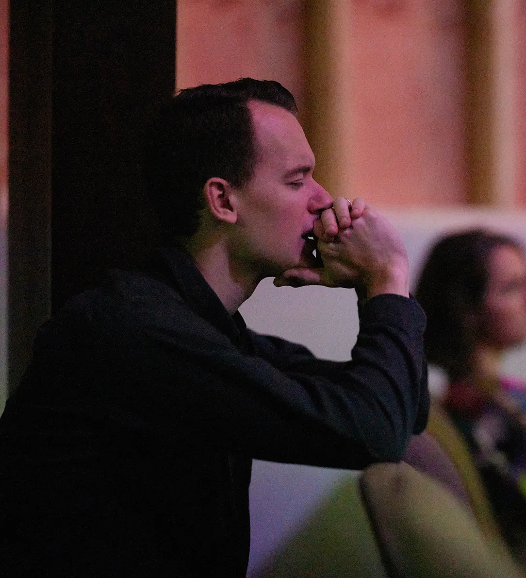 Man in dark clothing sitting thoughtfully with hands clasped near his mouth in a dimly lit room.