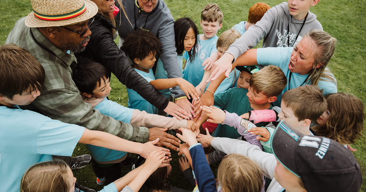 Group of children and adults standing in a circle outdoors with their hands stacked together in the center, showing teamwork and unity.