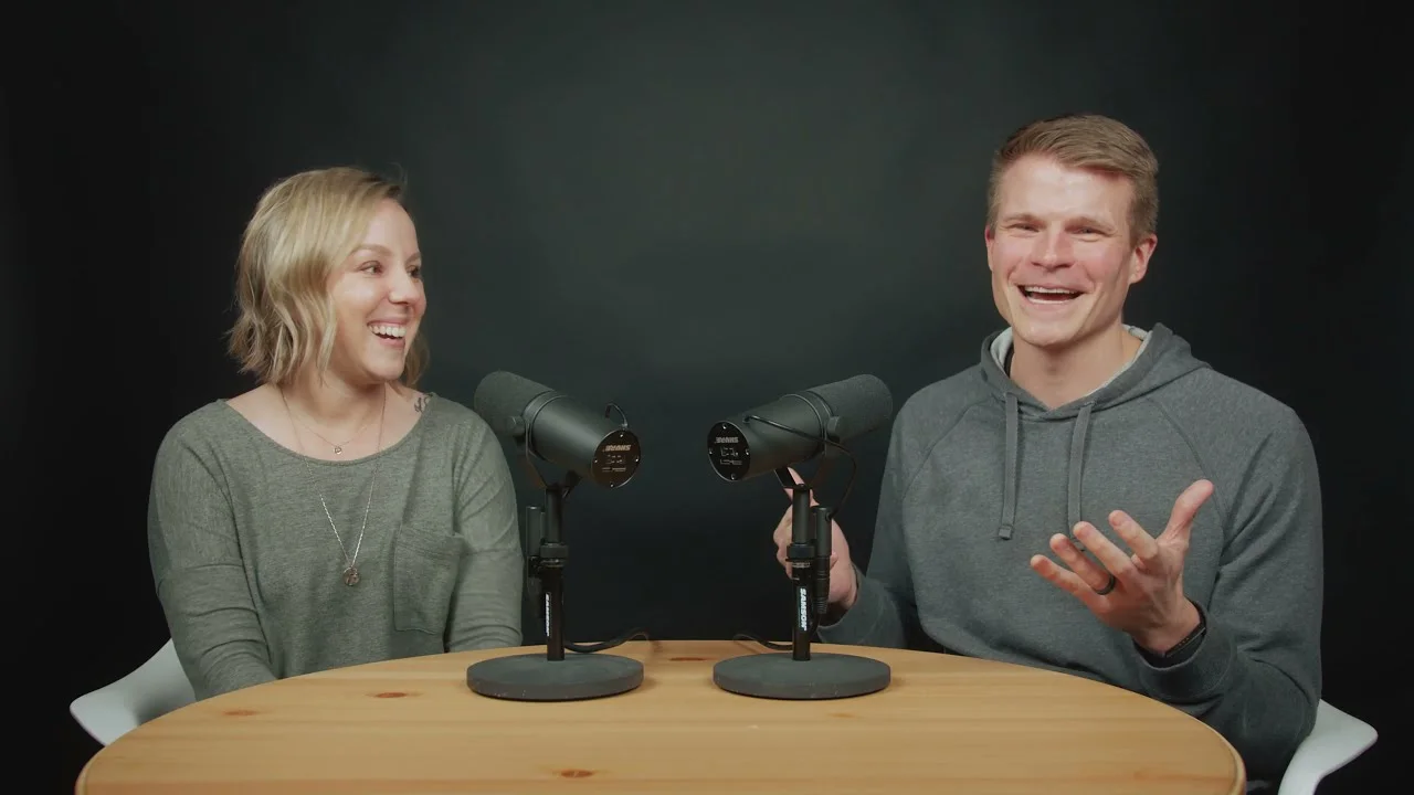Two people smiling and recording a podcast at a wooden table with microphones.