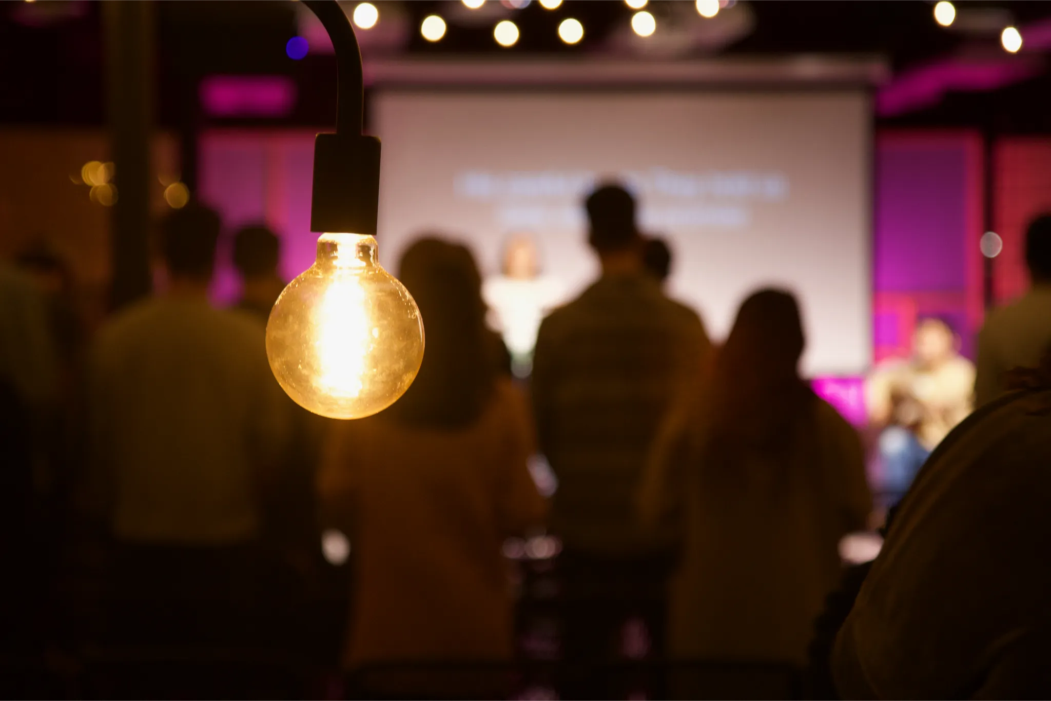 Glowing light bulb hanging in front of a blurred crowd at an indoor event.